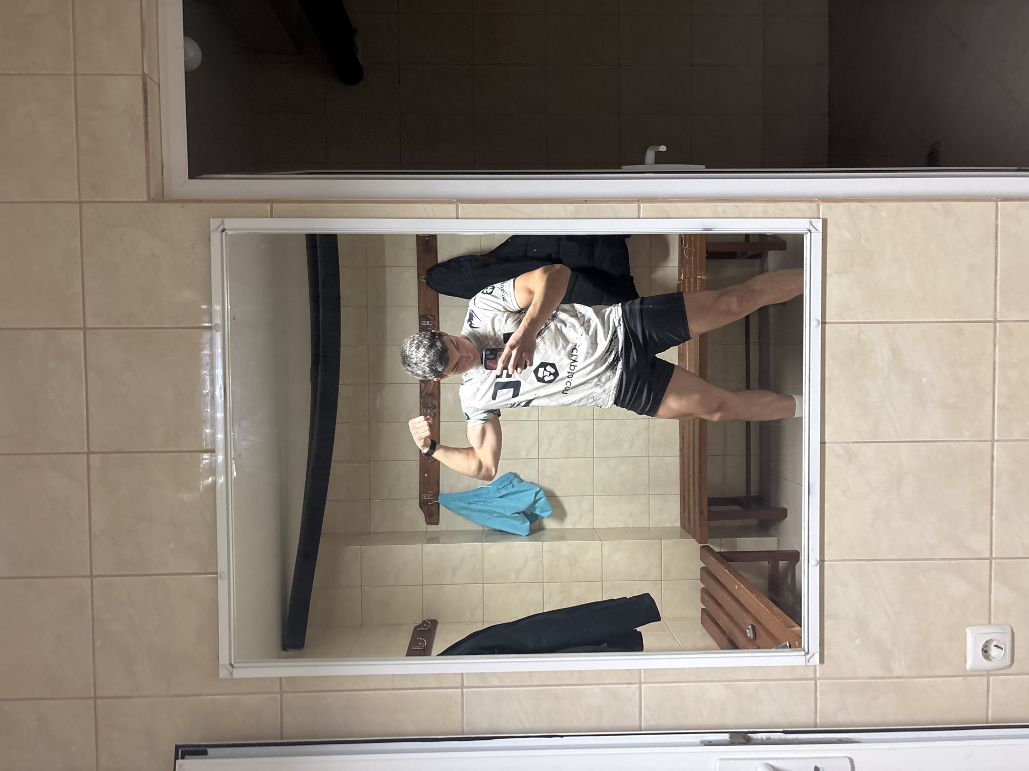 This image captures a mirror selfie of a man in what appears to be a changing room or locker room. The man, reflected in a large rectangular mirror with a white frame, is wearing a light-colored short-sleeved t-shirt with a graphic design and text (partially visible as "CLABDCU") and dark shorts. He is holding a smartphone in his right hand to take the photo and is flexing his left bicep, indicating he is posing. A watch or fitness tracker is visible on his left wrist.
The setting is characterized by beige or light brown tiled walls and floor, typical of a gym or sports facility changing area. Inside the mirror's reflection, behind the man, wooden benches or racks are visible, along with a black jacket draped over one and a pair of blue shorts or a towel on the floor. Above the mirror, a white frame, possibly of a door or window, borders a dark, indistinct background. To the right of the mirror, an electrical outlet is visible on the tiled wall. The scene is brightly lit, suggesting an indoor environment. Given the provided context, the photo was taken in Zlatograd, Bulgaria.