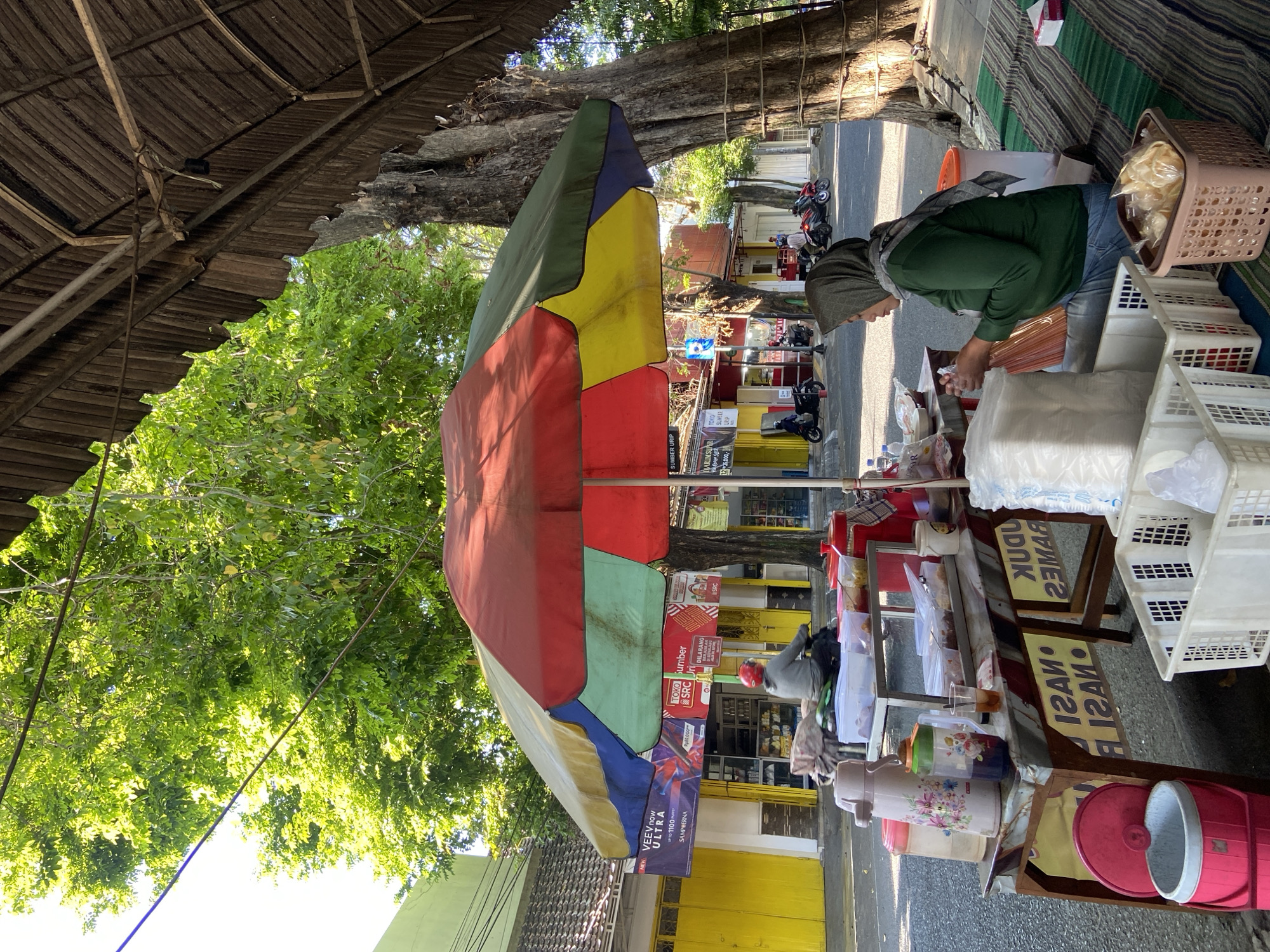 In Kecamatan Pati, Indonesia, a street food vendor is seen under a large, multi-colored umbrella. The vendor, a woman wearing a green hijab and green long-sleeved shirt, is seated on a striped mat, actively preparing food. In front of her, a wooden food cart displays various food containers, drinks in thermoses, and packaged goods. Behind the cart, the street is lined with shops, one of which has bright yellow doors and signage. Motorbikes are parked along the street, and a person on a motorbike is visible in the background. The environment appears to be a sunny day, with lush green trees providing shade.