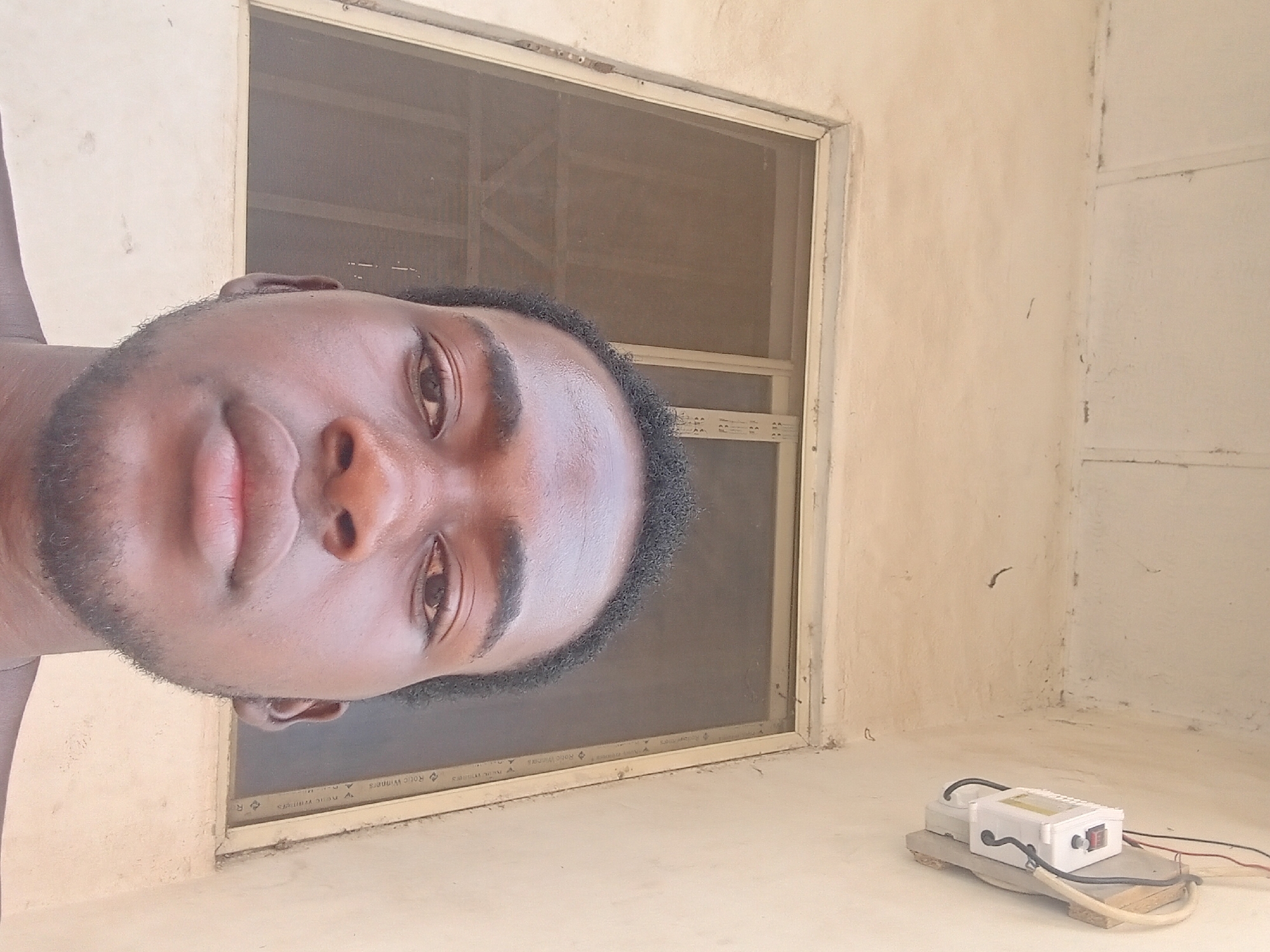 The image captures a close-up, horizontally oriented portrait of a young man, likely of African descent, indoors in Lokoja, Nigeria. He is positioned in the left half of the frame, looking towards the viewer with a neutral and calm expression. He has short, dark, curly hair and a light beard or stubble on his chin and cheeks. His eyes are brown, and his lips are full.
The background consists of light, off-white or beige-colored walls. Behind the man's head and extending to the right is a window with a mesh screen, possibly to keep out insects. The window frame appears to be made of light-colored material, possibly aluminum or painted wood. Faint text is visible on the lower part of the window frame, reading "Rolic Winners" repeated multiple times.
In the bottom right corner of the frame, placed on a slightly elevated surface or ledge of the same light-colored material as the walls, is a white electrical box. This box has a visible red power switch, and black and red wires are connected to it, suggesting it's a power supply, charger, or a DIY electrical setup. The box is resting on a small, light-colored wooden base.
The overall setting appears functional and simple, likely a room in a residence or basic structure. The natural light filtering through the window suggests it is daytime. The activity is a posed photograph, possibly a selfie given the close-up and slightly informal angle.