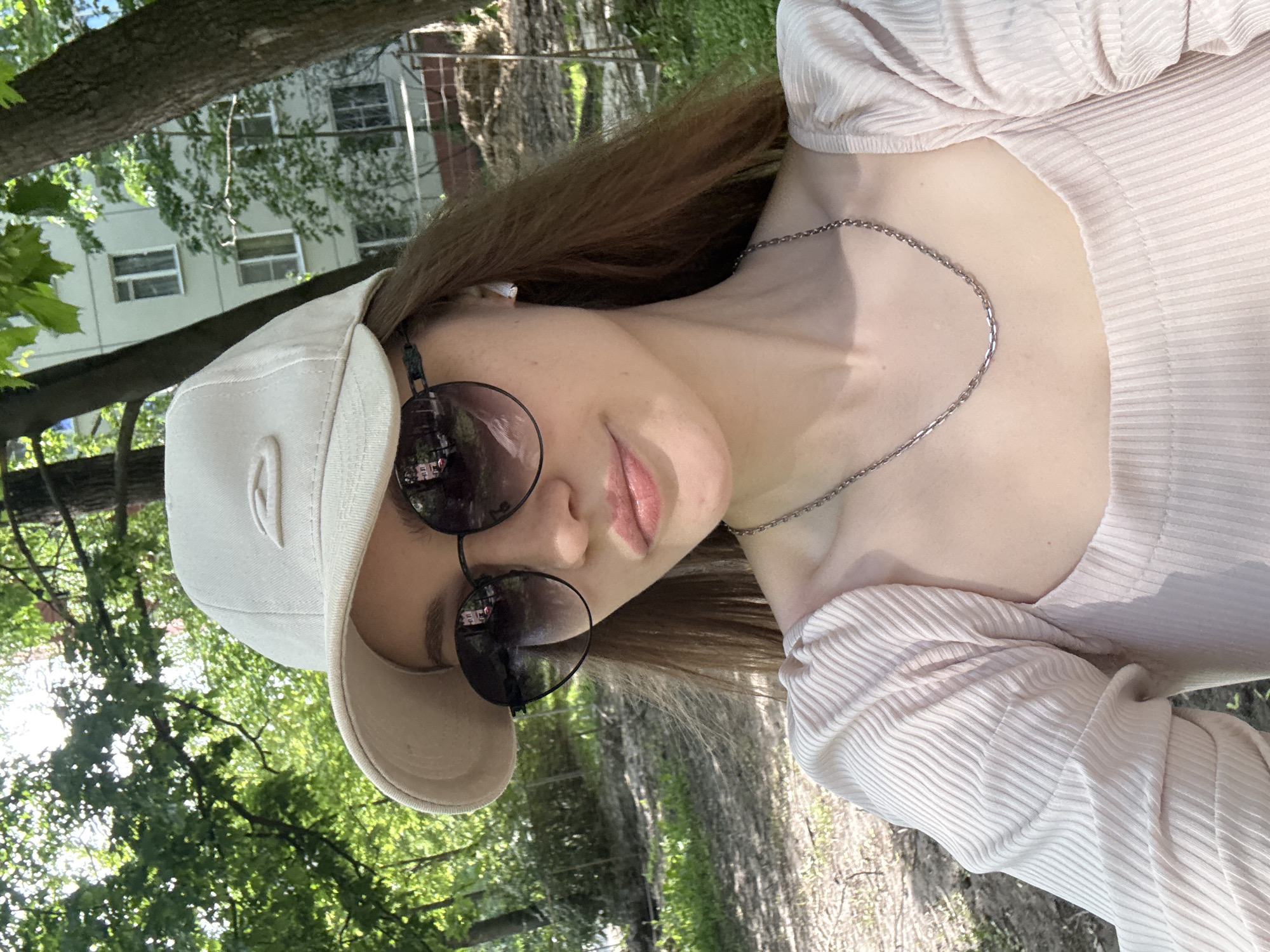 A young person, likely female, is featured in a close-up outdoor portrait, with the image rotated 90 degrees clockwise. She has long brown hair and is wearing a light beige baseball cap, dark round sunglasses, and a ribbed light-colored long-sleeve top. A thin silver-colored chain necklace is visible around her neck, and a white earbud is in her left ear. Her lips are slightly parted, and her expression is neutral, largely obscured by the sunglasses.
The background is filled with lush green foliage from trees, suggesting a park or a wooded area. Behind the trees, a light-colored multi-story building with visible windows can be seen. The scene is brightly lit by natural sunlight, indicating either midday or afternoon on a clear, sunny day. No specific activity beyond posing for the photograph is depicted. There is no visible text in the image.