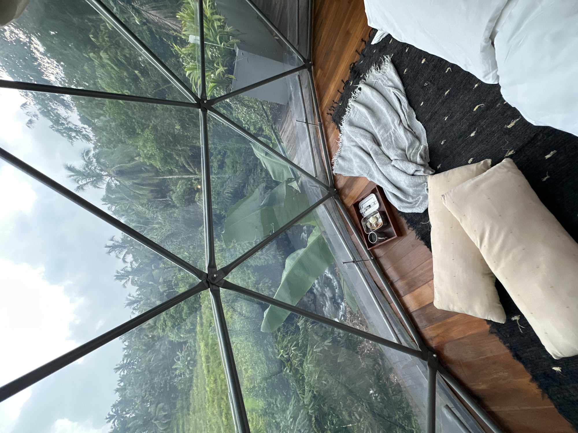 A high-angle shot inside a geodesic dome structure, likely a hotel room or lodging in Licin, Indonesia, overlooking a lush green landscape. The dome's frame is constructed of dark metal bars and transparent panels, providing a panoramic view of dense tropical foliage, possibly rice terraces, under an overcast sky.
Inside the dome, the scene is partially composed as a bedroom arrangement. A portion of a white bed with fluffy pillows and bedding is visible in the upper right corner, extending onto a black rug with light-colored embellishments. Near the edge of the bed is a light gray blanket, and a small dark brown wooden tray, containing a small plate with food and a coffee cup. Two large, light beige pillows rest on the edge of the rug. The flooring is dark brown wood.