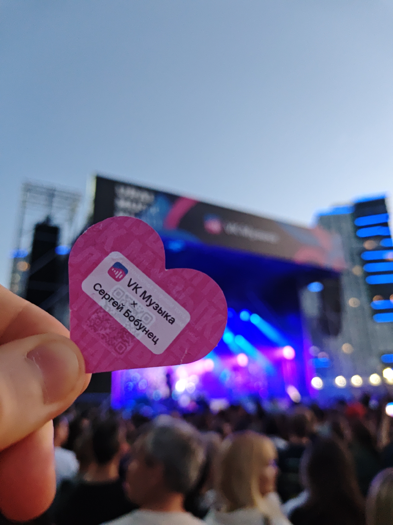 In a city park in Yekaterinburg, Russia, on a clear day, a person holds a pink heart-shaped sticker with Russian text and a QR code. The sticker reads "VK Музыка" and "Сергей Бобунец". In the background, a large outdoor concert stage with bright blue and pink lights is visible, with a blurred crowd of people in front of it. The overall scene suggests a music festival or event.