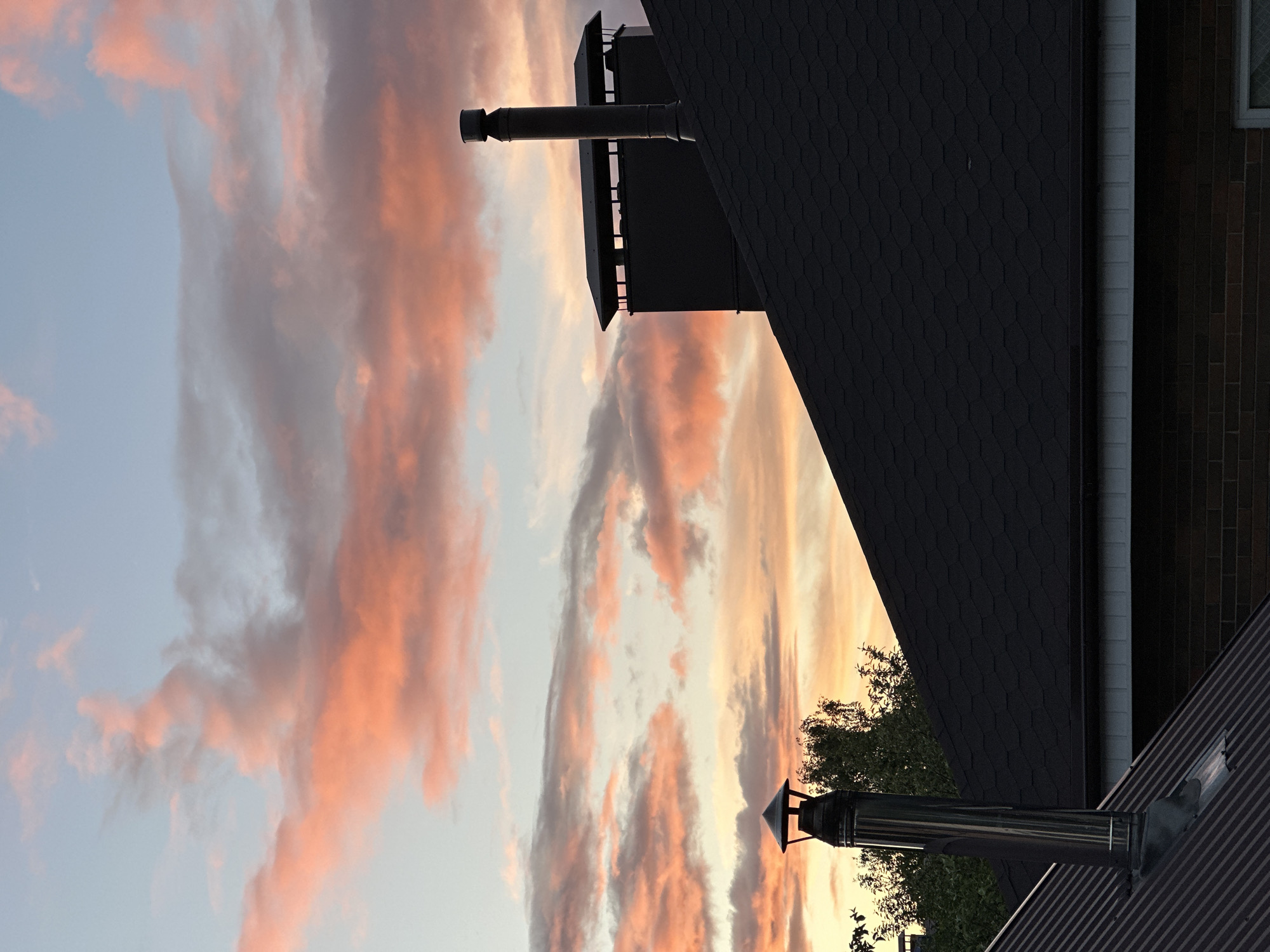 The image shows a partial view of buildings, chimneys, and a sky with clouds. The location in Nizhny Novgorod, Russia, is not explicitly depicted and is unknown.
Foreground: Two metallic chimneys are visible on rooftops. One is situated on a dark, corrugated roof, while the other is connected to a dark-colored building section with an accompanying structure.
Background: The sky is filled with clouds that have shades of peach and gray, suggesting a sunset or sunrise. A brick wall is seen on the right side of the image, and some greenery is vaguely visible between the chimneys.
Overall: The shot is taken from a low angle, looking up toward the buildings and the sky.