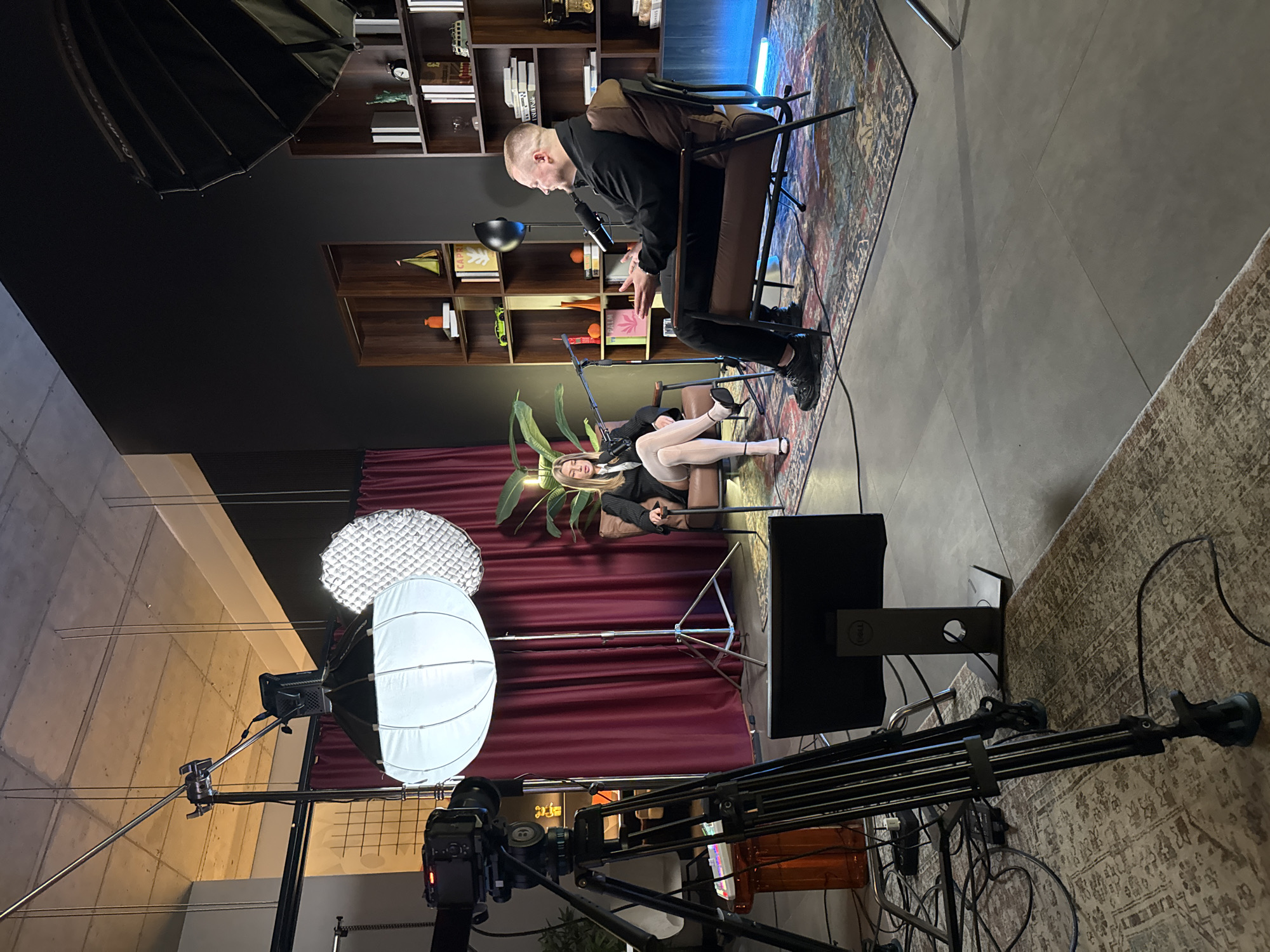 The image depicts a studio setting, likely located in Warsaw, Poland. A man in black attire sits on a chair facing a woman in a black suit and white stockings, also seated. They appear to be engaged in a conversation, possibly a podcast or interview. Behind them is a bookcase with decorative objects. Stage lighting equipment including two softboxes, is positioned in the foreground, along with a camera and tripod. The floor is carpeted with geometric patterns. In the background, a burgundy curtain is visible. A monitor with a DELL logo sits on a stand next to the woman.