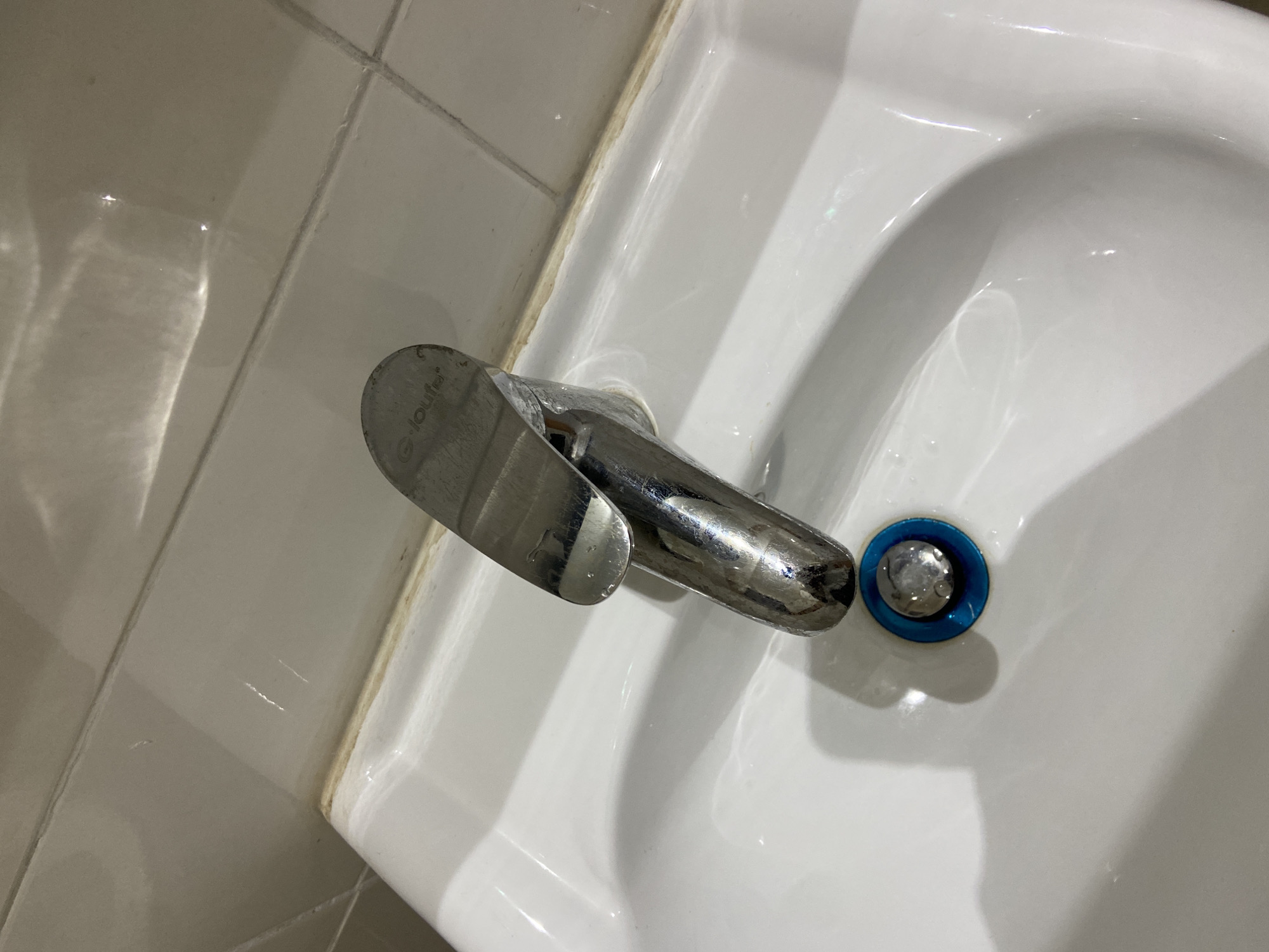 The image shows a close-up, overhead view of a white ceramic sink and a chrome faucet. The faucet has a lever handle with the logo "G-loufe" imprinted on it. Next to the faucet, there is a drain with a blue ring and a silver stopper. The sink is clean and appears to be in good condition. The background consists of white tiled walls, which are reflecting light from an unseen source. There are some water droplets and streaks visible on the sink and faucet, suggesting recent use. The lighting is bright, possibly from an overhead light or natural light coming from a window, creating strong reflections on the shiny surfaces. The overall scene is mundane, depicting a common bathroom or kitchen fixture. No people are visible in the image. The focus is entirely on the sink and faucet.