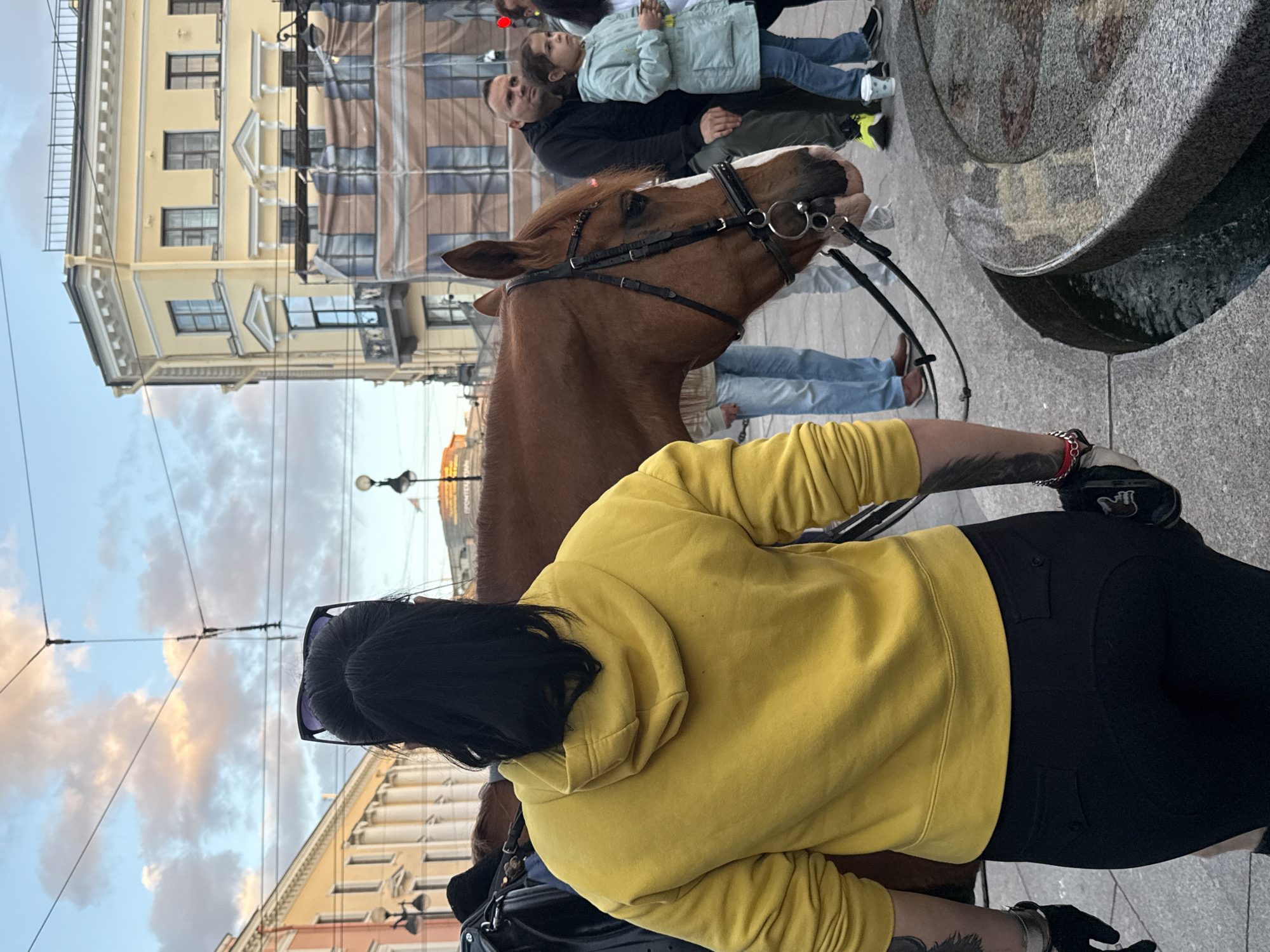 An outdoor daytime scene in Saint Petersburg, Russia, features a brown horse with a black bridle standing near a stone fountain. The horse's head is lowered, facing towards the right of the frame. Behind the horse, a woman with black hair pulled up and sunglasses on her head, wearing a yellow hooded sweatshirt and black pants, is seen from the back. To the right of the horse, a man in a black hooded jacket and a young girl in a light blue jacket are visible. The background shows architectural elements characteristic of Saint Petersburg, including a yellow building with columns and elaborate window frames, and a building undergoing renovation or covered with scaffolding and tarps. Overhead, power lines and a cloudy sky are present. The ground is paved with cobblestones and stone.