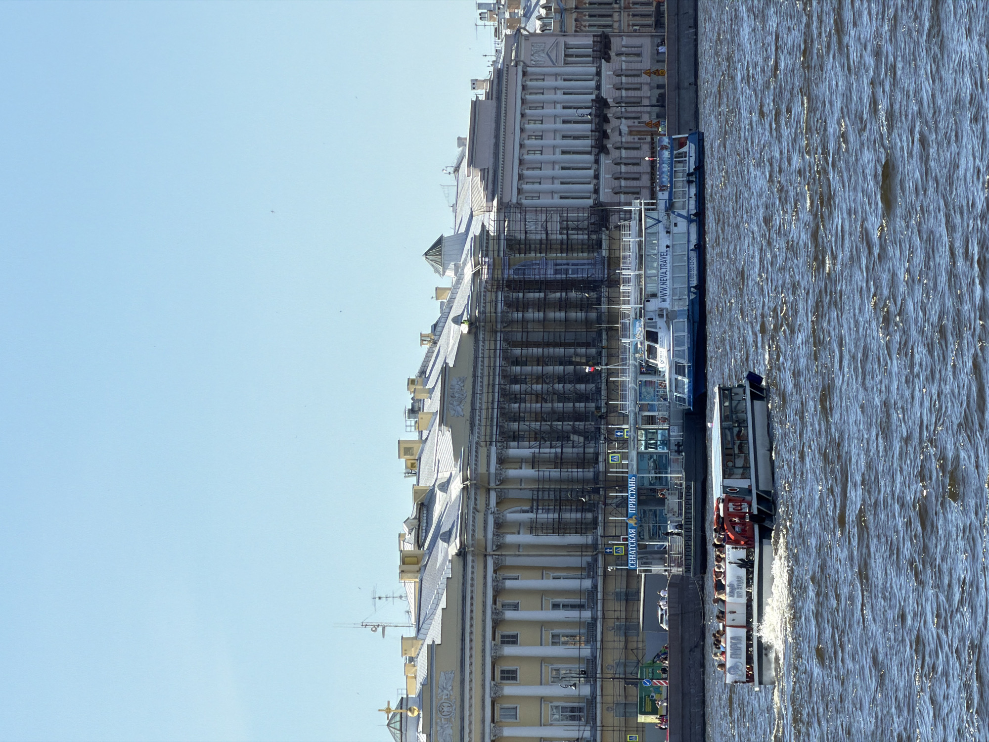 The image depicts a sunny day on the Neva River in Saint Petersburg, Russia, with the Senate and Synod Building prominently featured in the background. The building, undergoing renovations with scaffolding visible, displays a grand Neoclassical facade with tall columns and a yellow stucco finish. A large tour boat carrying passengers navigates the choppy, dark blue water in the foreground. Several other vessels are docked or moored near a pier labeled "СЕНАТСКАЯ ПРИСТАНЬ" (Senate Pier) on the right side of the frame. The sky is a clear, pale blue.