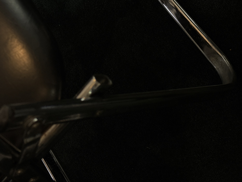 The image provides a close-up, dimly lit view of a segment of an object, consistent with a piece of modern furniture. Prominent features include reflective, tubular metal components, likely chrome-plated or polished steel. A vertical metal bar is visible on the left side, connected horizontally to a shorter, circular tube extending towards the right. In the lower right quadrant, a curved, L-shaped metal element, consistent with a furniture base, is present. The majority of the scene consists of a dark, textured surface, resembling a carpet or fabric upholstery. In the upper right, a dark, curved surface, possibly a seat or armrest constructed from leather or dark plastic, displays a diffused, warm reflection. No individuals, actions, or interactions are present. The environment is an interior space with limited illumination. The specific location within Mexico City, Mexico, cannot be ascertained from the visual information provided.