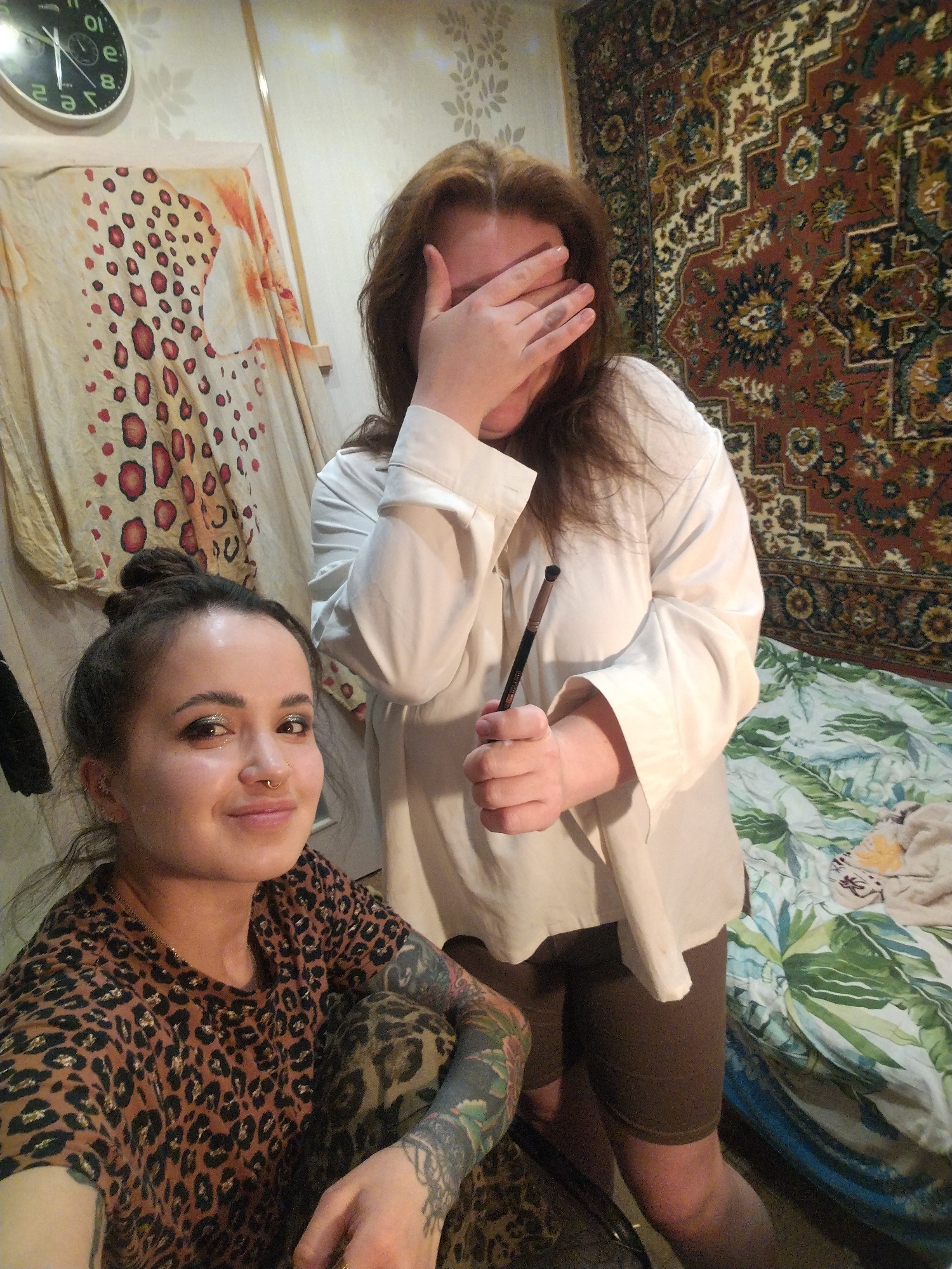 An indoor scene in a private residence located in Unknown, Unknown, features two adult females. On the left and foreground, one female with dark hair pulled into a bun and a septum piercing smiles directly at the camera. She wears a brown and black leopard print t-shirt and displays extensive dark tattoos on her left arm. Glitter eyeshadow is visible on her eyelids. Behind her and to the right, a second female with long, reddish-brown hair covers her face with her right hand, obscuring her eyes. She wears a loose white long-sleeved shirt and dark brown shorts, holding a dark-handled makeup brush in her left hand. The background includes a light-colored fabric with red and black spots hanging on the left wall, a partially visible wall clock with inverted numbers, and a dark, ornate tapestry with a red, blue, and gold geometric pattern hanging on the right wall. A bed with a blue and green tropical leaf-patterned comforter is visible in the lower right background. The lighting suggests an artificial source.