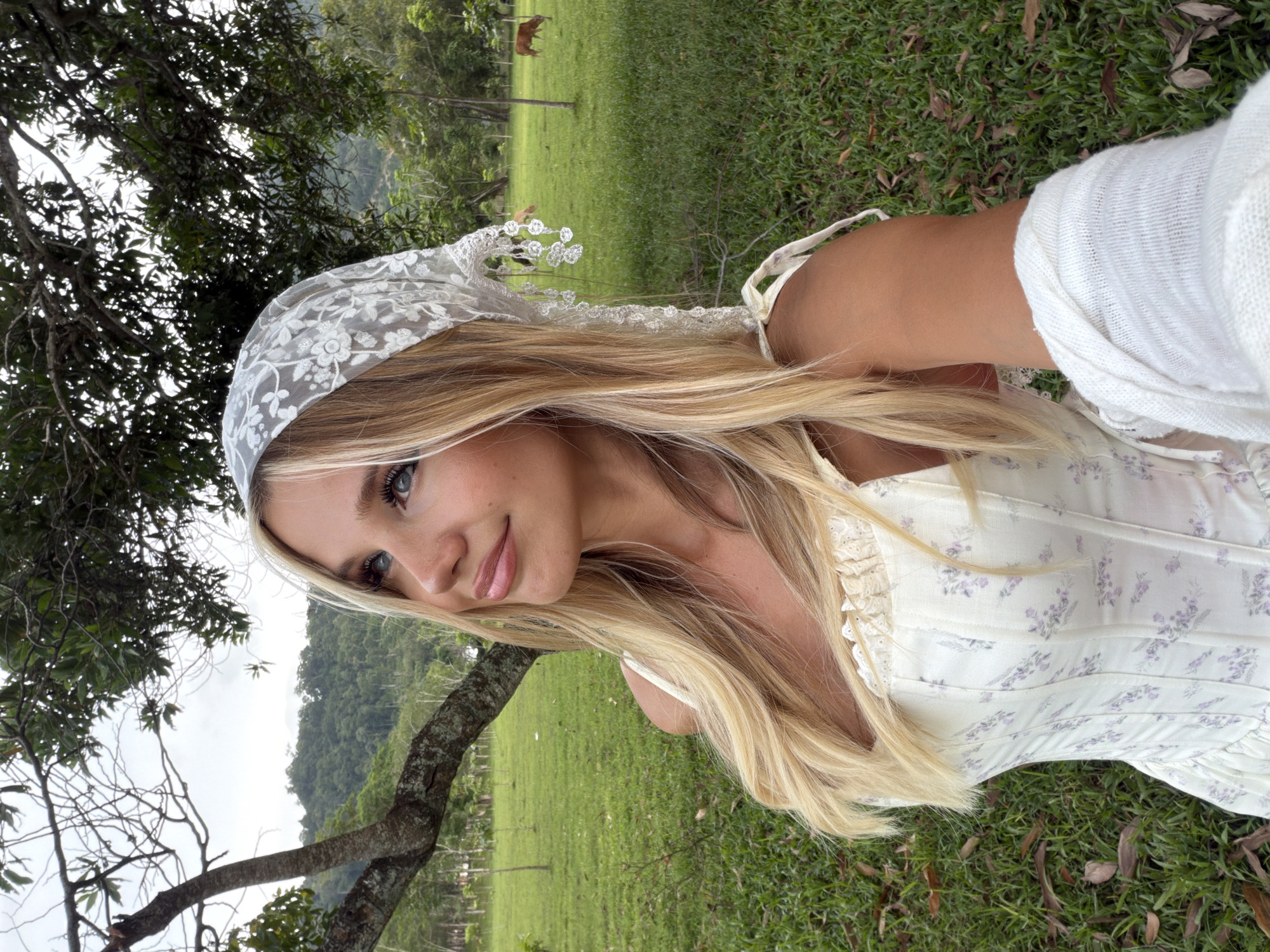 A young blonde woman with blue eyes, wearing a white floral corset and a white lace headscarf, poses for a selfie outdoors in a grassy field. She is looking upwards and to her left, with a slight smile. The background features a large tree trunk on the left, a lush green field, and a distant line of trees and hills, suggesting a rural or park-like setting. Two cows are visible grazing in the field in the background. The image appears to be taken in a warm climate, possibly during the day, with overcast skies. The woman's arm is extended, holding what appears to be a phone, capturing the selfie. The specific location in Ko Kaeo, Thailand cannot be determined from the visual information present in the image alone.