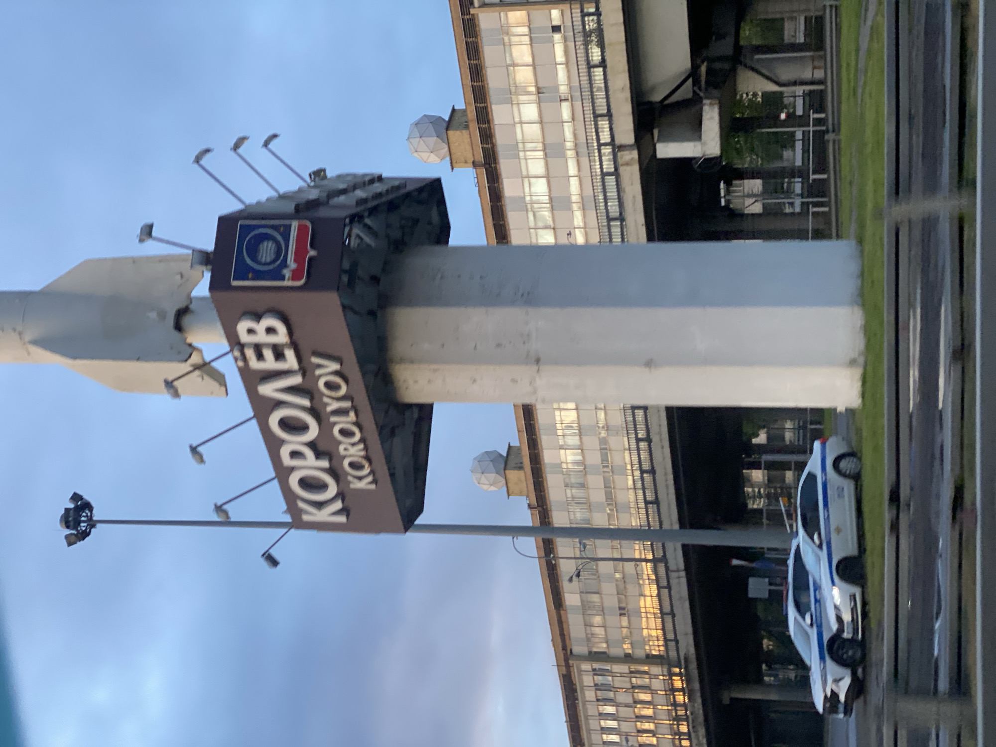 The image shows a sign in the city of Korolyov, Russia. The sign features the city's name in both Cyrillic ("КОРОЛЁВ") and Latin ("KOROLYOV") alphabets. Above the name is an emblem of the city, next to a rocket-shaped structure. The sign is illuminated by several lights extending from its structure. A large, hexagonal pillar supports the sign. In the background is a long building with a white facade and many windows. In the foreground, two white and blue vehicles are parked on the side of the road. The sky above is light blue.