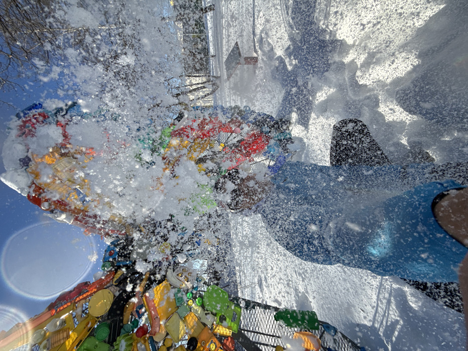 The image depicts a snowy outdoor scene in Gorky Park (Central Park of Culture and Leisure named after Maxim Gorky) in Kazan, Russia. A person, wearing a blue jacket and dark pants, is partially obscured in the foreground by a dense burst of white snow particles, appearing to be in a crouching or kneeling position. To the left, a large, intricate sculpture constructed from numerous multicolored recycled plastic objects (such as bottles and containers) is visible, partially covered in snow. The sculpture exhibits a complex, organic form with varied hues. The ground is uniformly covered in snow, and bare deciduous trees are present in the background. A small, red-topped stand, possibly a solar panel or information display, is also visible in the snowy expanse to the right. The sky is bright blue, and lens flare is present, indicating a sunny winter day.