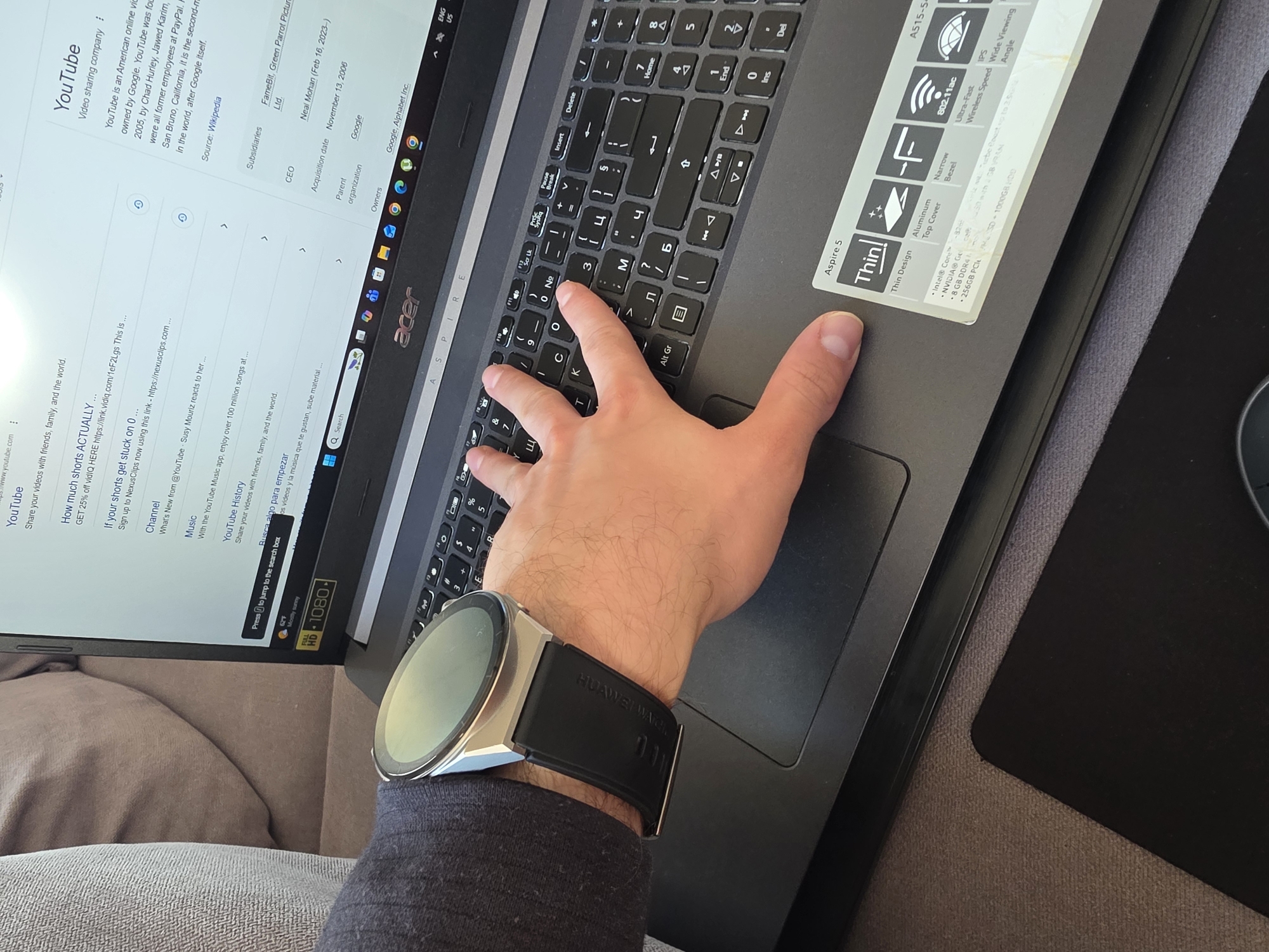 The image depicts a close-up view of a person using a laptop. The person's hand is resting on the keyboard, with a silver smartwatch with a black band visible on their wrist. The laptop is an Acer Aspire 5. The screen displays a YouTube page with various video suggestions and information about YouTube as a company. The laptop is resting on a gray surface, possibly a couch or other soft furnishing, alongside a black mousepad. There is no indication of the location in Yagodovo, Bulgaria.