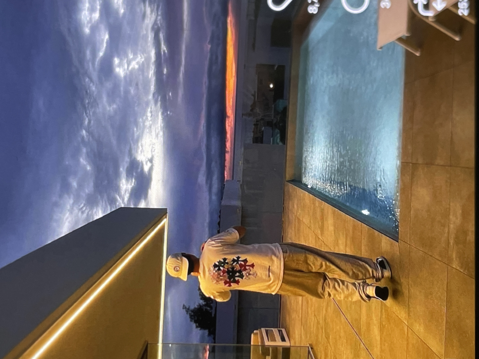 The image shows a person standing on a tiled patio next to an illuminated swimming pool. The person is wearing a white t-shirt with a graphic design on the back, light-colored pants, and a baseball cap. They are facing away from the camera, looking towards a dramatic sunset over a body of water. The sky is filled with colorful clouds, ranging from deep purple to fiery orange. In the background, there are structures that appear to be part of a modern building complex, possibly a rooftop terrace. The lighting suggests it is either dusk or dawn. The overall scene conveys a sense of relaxation and appreciation of nature's beauty.