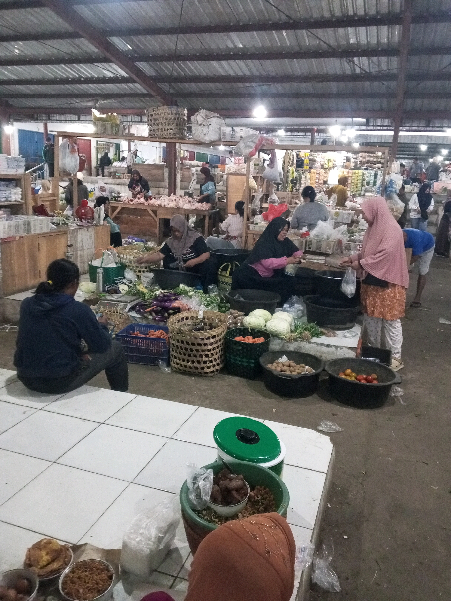The image shows an indoor market scene in Gentan, Indonesia, with numerous people and a variety of goods. In the foreground, a person wearing a dark blue hoodie and gray pants sits on a tiled floor, facing away from the camera. To their right, a variety of vegetables, including eggplants, carrots, and cabbages, are displayed in baskets and crates. A woman wearing a black hijab and pink shirt is seated behind a stall, interacting with another woman in a pink hijab and patterned skirt, who is holding a plastic bag. Several large black bowls hold produce such as potatoes and tomatoes. In the background, stalls are set up with more goods, including what appear to be chickens on display. The market has a corrugated metal roof supported by exposed beams. The overall environment is bustling, with shoppers and vendors engaged in commerce.