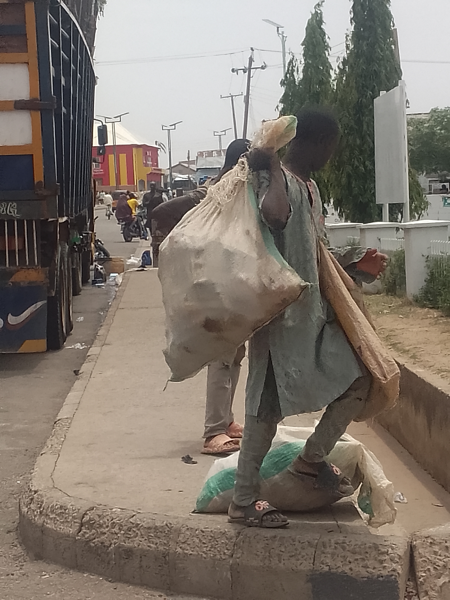 In Pantami, Nigeria, a child stands on the edge of a concrete sidewalk beside a road, carrying a large sack over their shoulder and another on their foot. The child wears worn clothing and sandals. Nearby, another individual is walking. In the background, there is a road with motorcycles and pedestrians, a colorful building, power lines, and tall green trees. A large truck occupies part of the left side of the frame.
