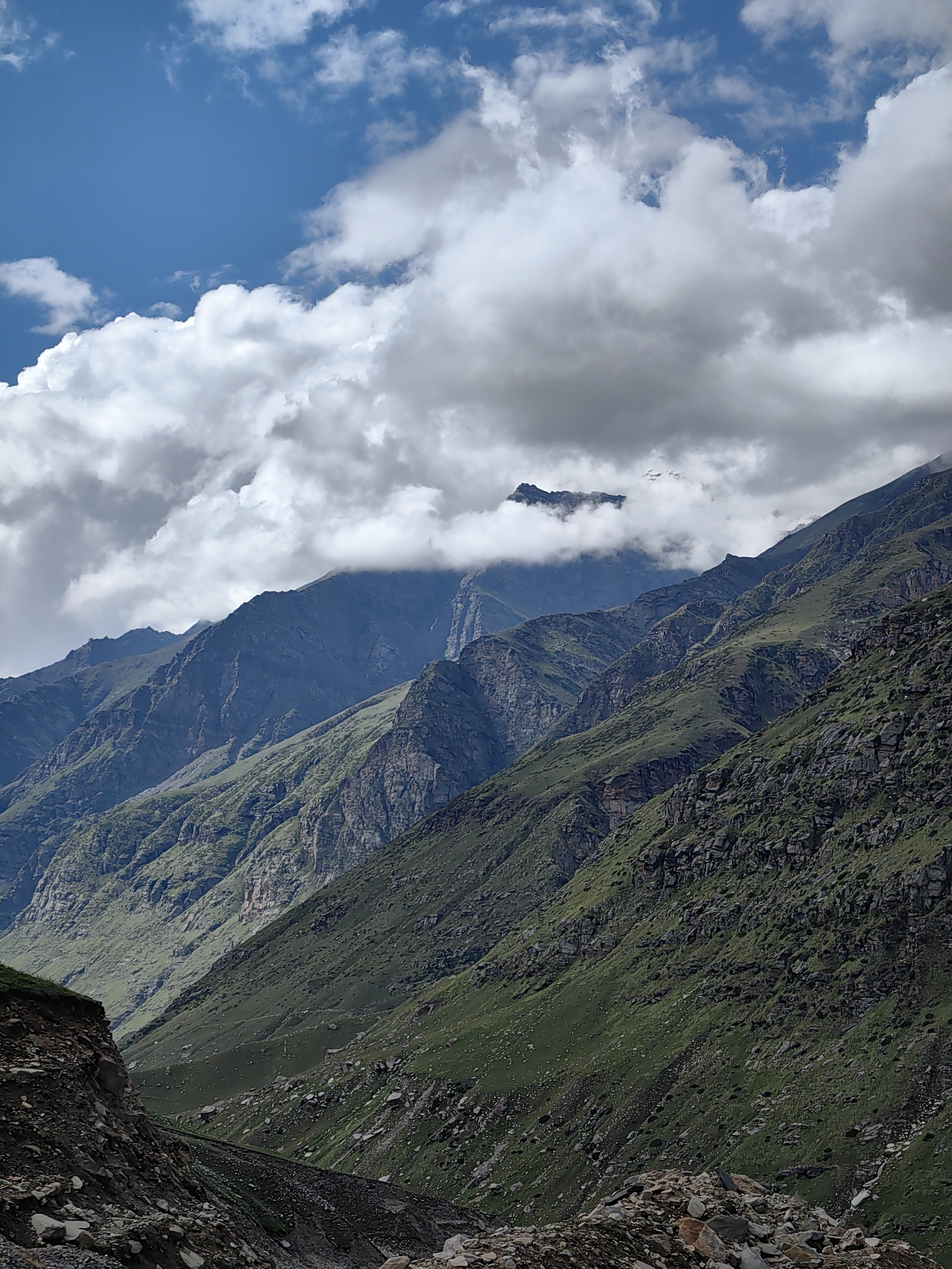 The image depicts a dramatic mountain landscape under a partially cloudy sky. In the foreground, the lower slopes of the mountains are covered in green vegetation and rocks. As the eye moves up, the terrain becomes steeper and rockier, with jagged peaks disappearing into thick, white clouds. The sky above is a vibrant blue, with large, puffy clouds casting shadows on the mountains below. The overall impression is one of raw, untamed natural beauty, characteristic of the higher altitudes of India. The location is not specified, but the scene suggests a remote, mountainous region.