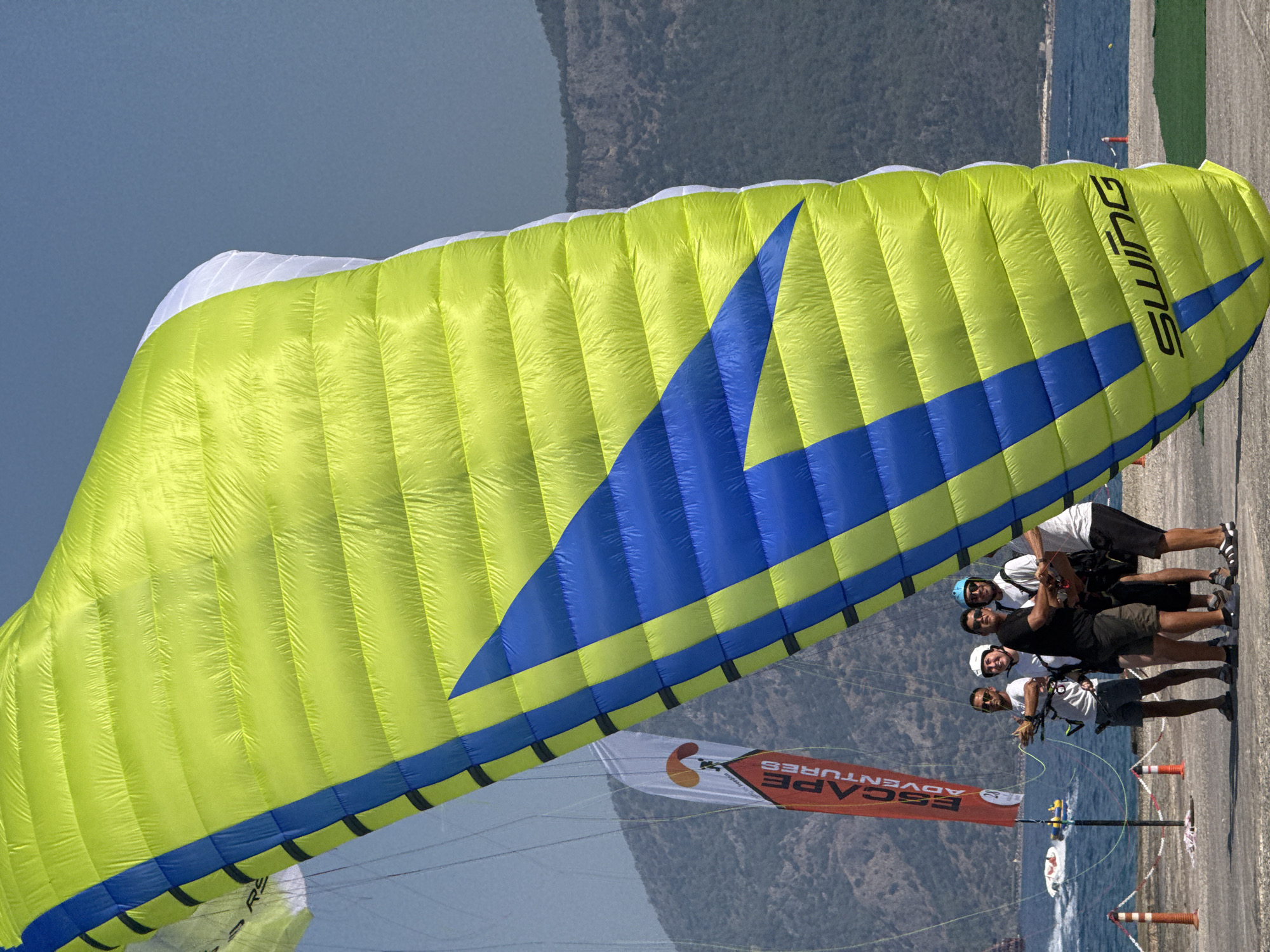 In Fethiye, Turkey, four men stand near the water, preparing for paragliding. A large, bright green and blue paraglider occupies the foreground, its canopy partially inflated. To the left, a red banner with white lettering reads "ESCAPE ADVENTURES." In the background, calm water stretches towards a distant, tree-covered mountain range. Several boats are visible on the water. The ground is paved, with orange cones and white ropes marking a boundary. The sky is clear and blue.