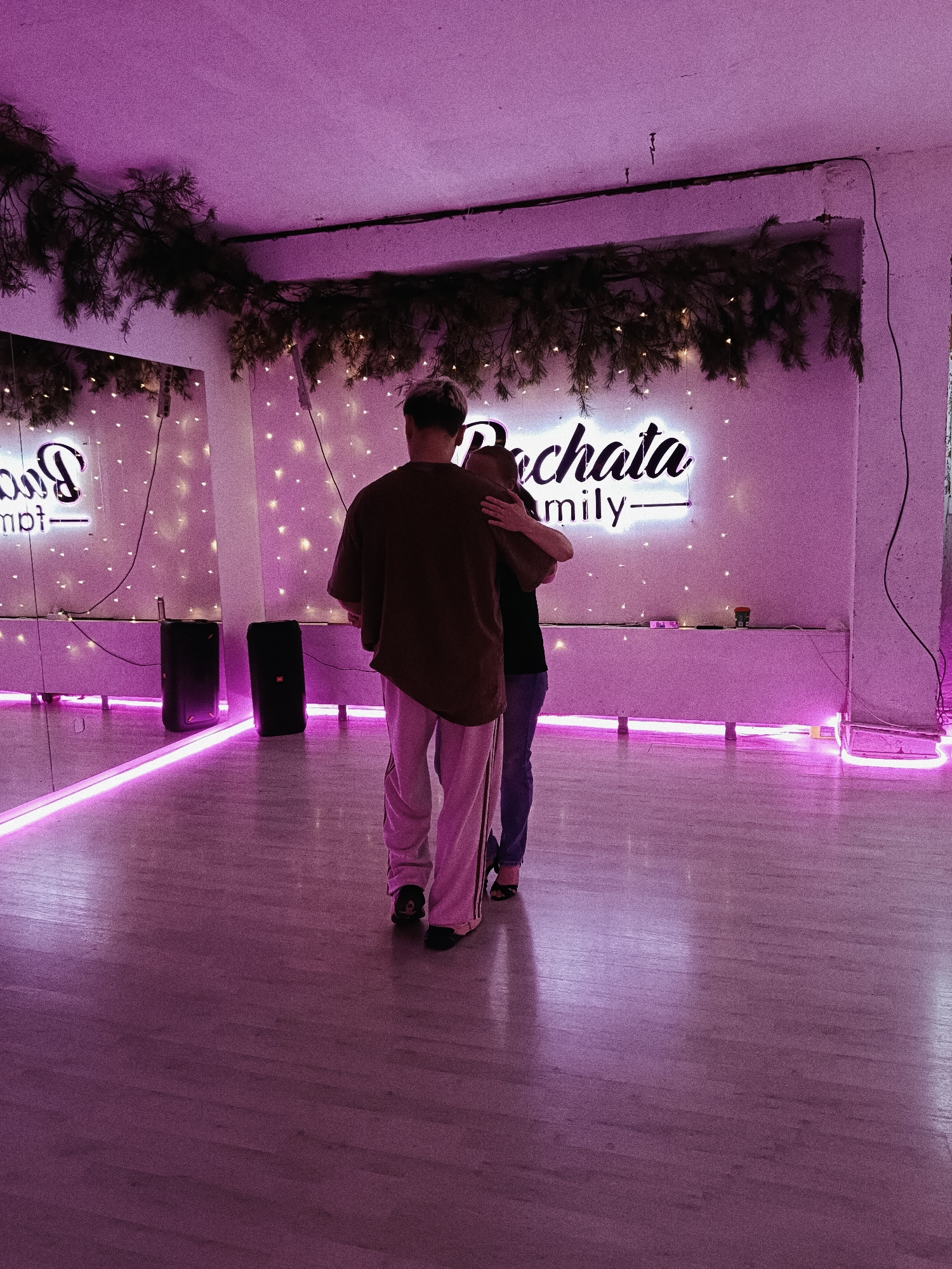 This image depicts an indoor scene, likely a dance studio or event space, in Chaikovsky, Russia, bathed in strong purple and pink lighting. Two individuals, viewed from behind, are the central subjects, appearing to be in a close embrace or dancing. The person on the left wears a brown oversized t-shirt and light-colored, possibly pinkish, loose-fitting pants, with short, light-toned hair. The second person, partially obscured behind the first, wears dark clothing and high-heeled shoes.
The room features a light-colored wooden floor and pale walls, heavily influenced by the purple and pink LED lights. A prominent neon sign on the back wall spells out "Bachata Family." Above this sign, a decorative garland of evergreen branches, possibly pine or fir, is strung with small, warm lights.
On the left side of the room, a large mirror reflects parts of the space and the "Bachata Family" sign (appearing as "Bao mpt-"). Two dark, rectangular speakers stand on the floor near the mirror. Pink LED strips are visible along the base of the walls and possibly embedded in the floor, contributing to the distinctive lighting scheme. The setting suggests an evening event, likely a dance class or social gathering focused on partner dancing, given the "Bachata" text.