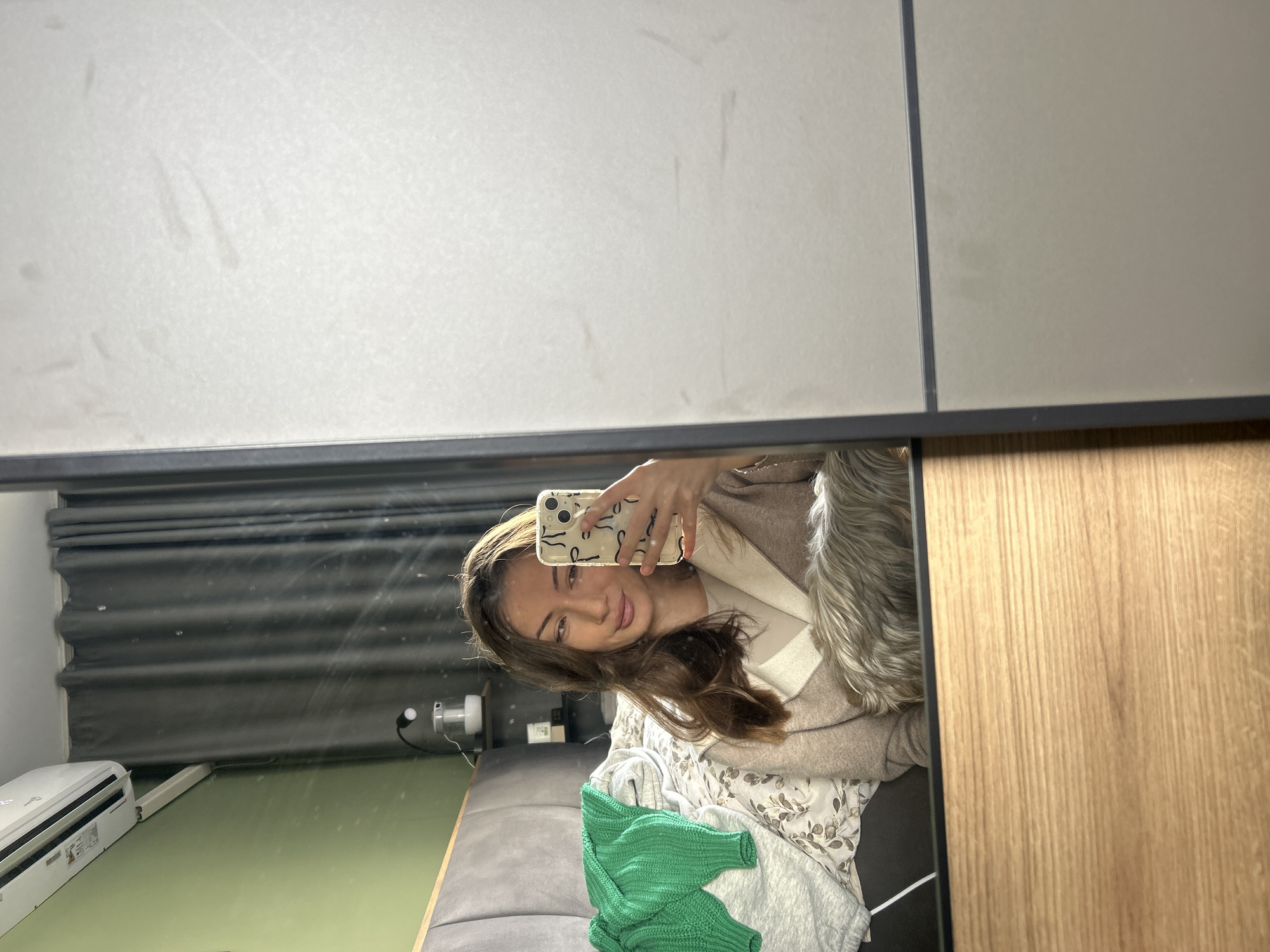 The image shows a selfie taken in a mirror. The reflection reveals a young woman holding a smartphone. She has brown hair and is wearing a beige coat over a white top. There is a green sweater on a bed. The room has a gray bed, green floor, gray curtains, and an air conditioner unit. The mirror is attached to a wardrobe with wooden-patterned doors. The location is in Burgas, Bulgaria.