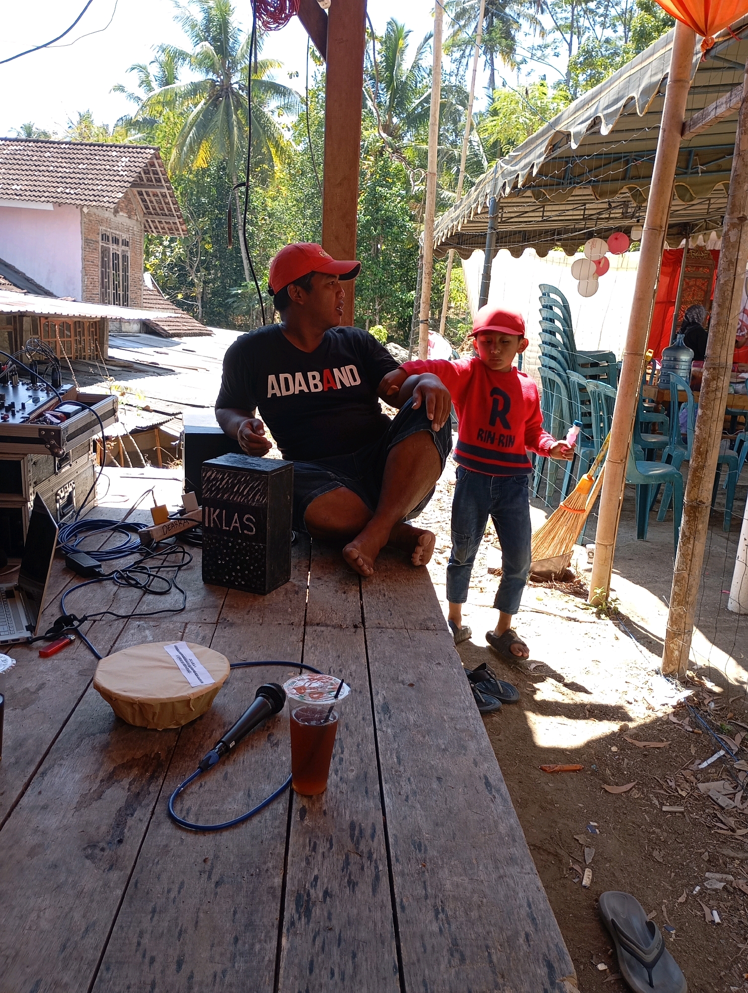 An outdoor scene at the Ketepung, Indonesia, shows a man and a child sitting at a wooden table. The man, wearing a red cap and a black t-shirt with the word "ADABAND" printed on it, is seated on the right. A child, also wearing a red cap and a red sweater with the text "RIN RIN" visible, stands to the left of the man. On the table in front of them, there is a microphone, a cup of iced tea, and a round object wrapped in paper. Behind them, a canopy is erected, with chairs stacked underneath. The environment appears to be a rural or semi-rural area, with trees and buildings visible in the background. Sunlight casts shadows on the ground, suggesting it is daytime.