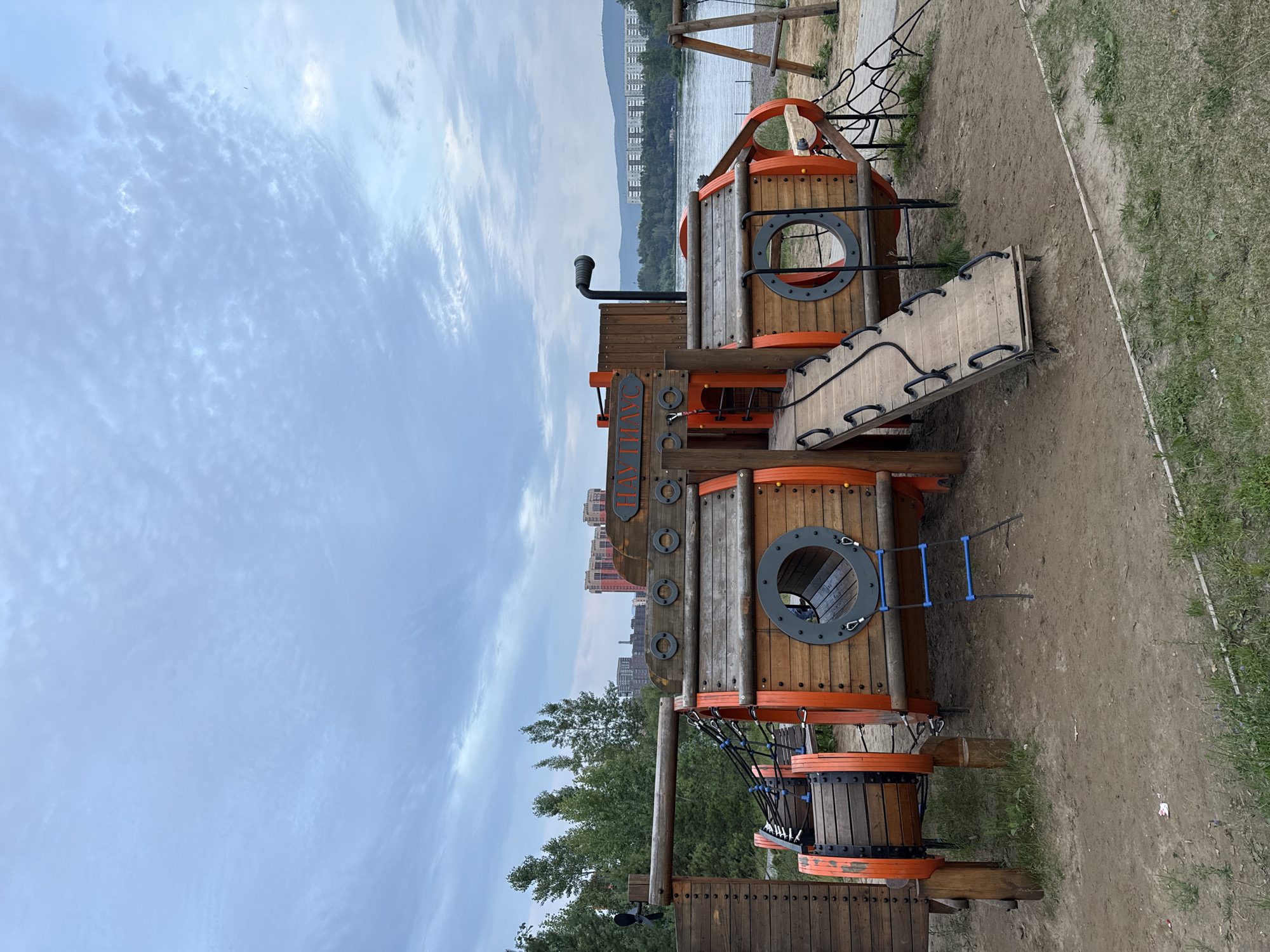 The image shows a wooden playground structure designed to resemble a ship, located in a park next to a body of water, likely the Yenisei River, in Krasnoyarsk, Russia. The playground features wooden beams, circular openings that serve as windows or entrances, and a ramp with handrails. A ladder with blue rungs is attached to the side of the structure. In the background, there are trees, buildings, and a cloudy sky. The ground in front of the playground is sandy and uneven, with patches of grass. There are no people visible in the image. The overall scene depicts an outdoor recreational area.