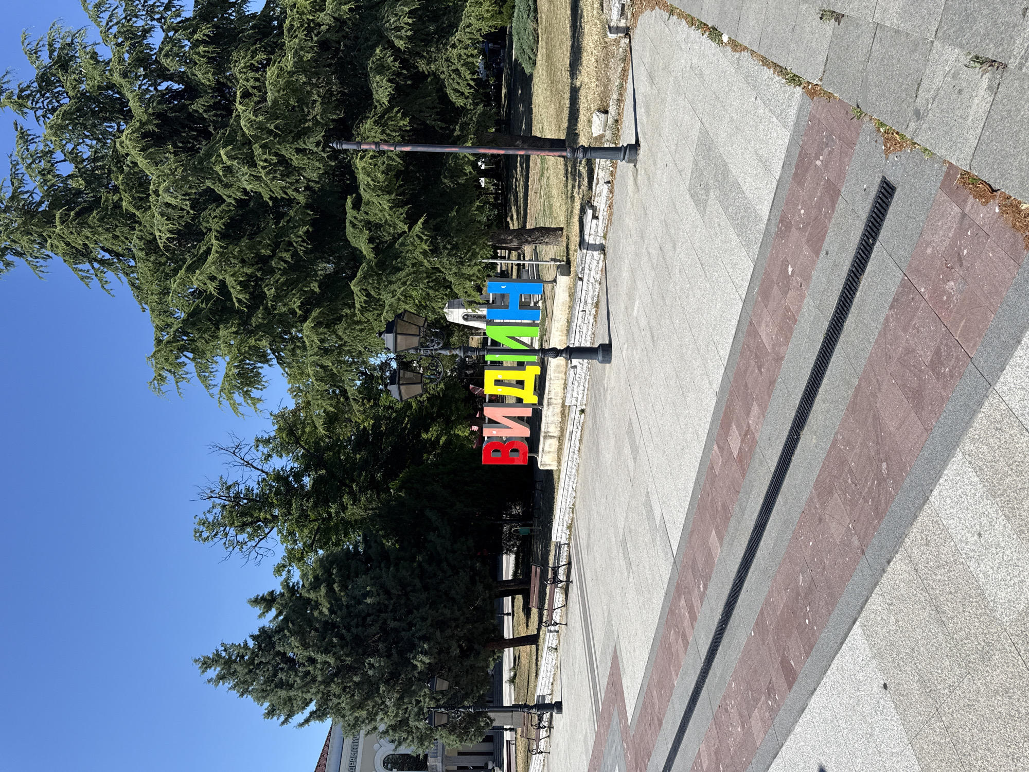The image shows a street scene in Vidin, Bulgaria. In the center of the frame, large, colorful letters spell out "VIDIN" in red, yellow, blue, and green. The letters are three-dimensional and appear to be made of metal. They are positioned in front of a lush green tree and a clear blue sky. The letters are arranged in a vertical line, with each letter standing upright.
To the left of the letters, there are tall, mature evergreen trees with dense foliage. A lamppost with an ornate design stands in front of the letters. In the background, there are more trees and some buildings, including a white building with an arched entrance. To the right of the letters, a paved walkway made of light-colored stone tiles with red accents is visible. A dark metal grate runs along the edge of the walkway. There are also benches visible, suggesting a public seating area.
The sun is shining brightly, casting shadows on the ground and indicating it is daytime. The weather appears to be clear and sunny. The overall scene suggests a public space, possibly a park or a plaza, in a town or city. The prominent display of the town's name implies it's a landmark or a point of interest. No people are visible in the image. The image is taken from a slightly elevated angle, looking down on the scene. The overall mood is bright and cheerful, enhanced by the vibrant colors of the letters.