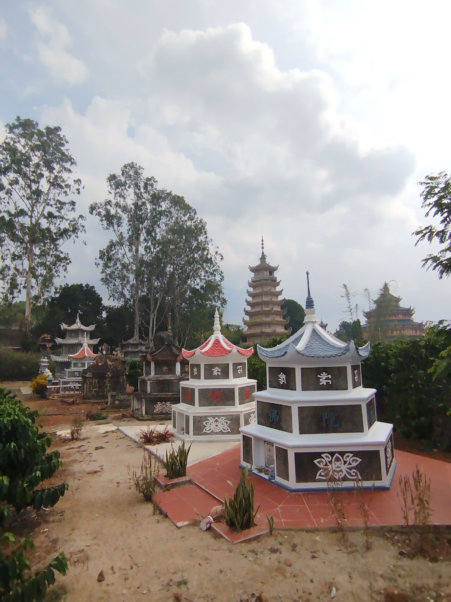 The image captures a solemn and serene landscape in Phú An, Vietnam, likely a memorial garden or cemetery associated with a Buddhist temple. In the foreground, several elaborately decorated, multi-tiered memorial structures or stupas are visible. The closest two are prominent, featuring white bases, dark gray middle sections, and contrasting roofs – one with a red roof, the other with a blue-gray roof. These structures are adorned with intricate white floral or cloud-like patterns and visible text in what appears to be Chinese or Vietnamese Han characters. For example, characters like "佛" (fú, Buddha), "陀" (tuó, Buddha), and "念" (niàn, remember/chant) are discernible, along with Buddhist symbols such as the swastika (卍), signifying good fortune. Small potted plants and dry foliage are placed around their tiled platforms.
The ground is a mix of dirt paths and reddish-brown tiles, suggesting a well-maintained but rustic environment. Tall, slender trees with sparse foliage, possibly eucalyptus, punctuate the midground, with lush green bushes and smaller plants filling the spaces between the memorial structures.
In the background, partially obscured by trees, stand two larger, multi-story pagodas or temple buildings, echoing the architectural style of the foreground structures. One is a tall, slender pagoda with multiple tiers, while the other appears broader and more ornate.
The scene is set under a bright, cloudy sky, indicating daytime, possibly mid-morning or early afternoon, with soft, diffused light. No people are visible, contributing to the tranquil and contemplative atmosphere of the site.