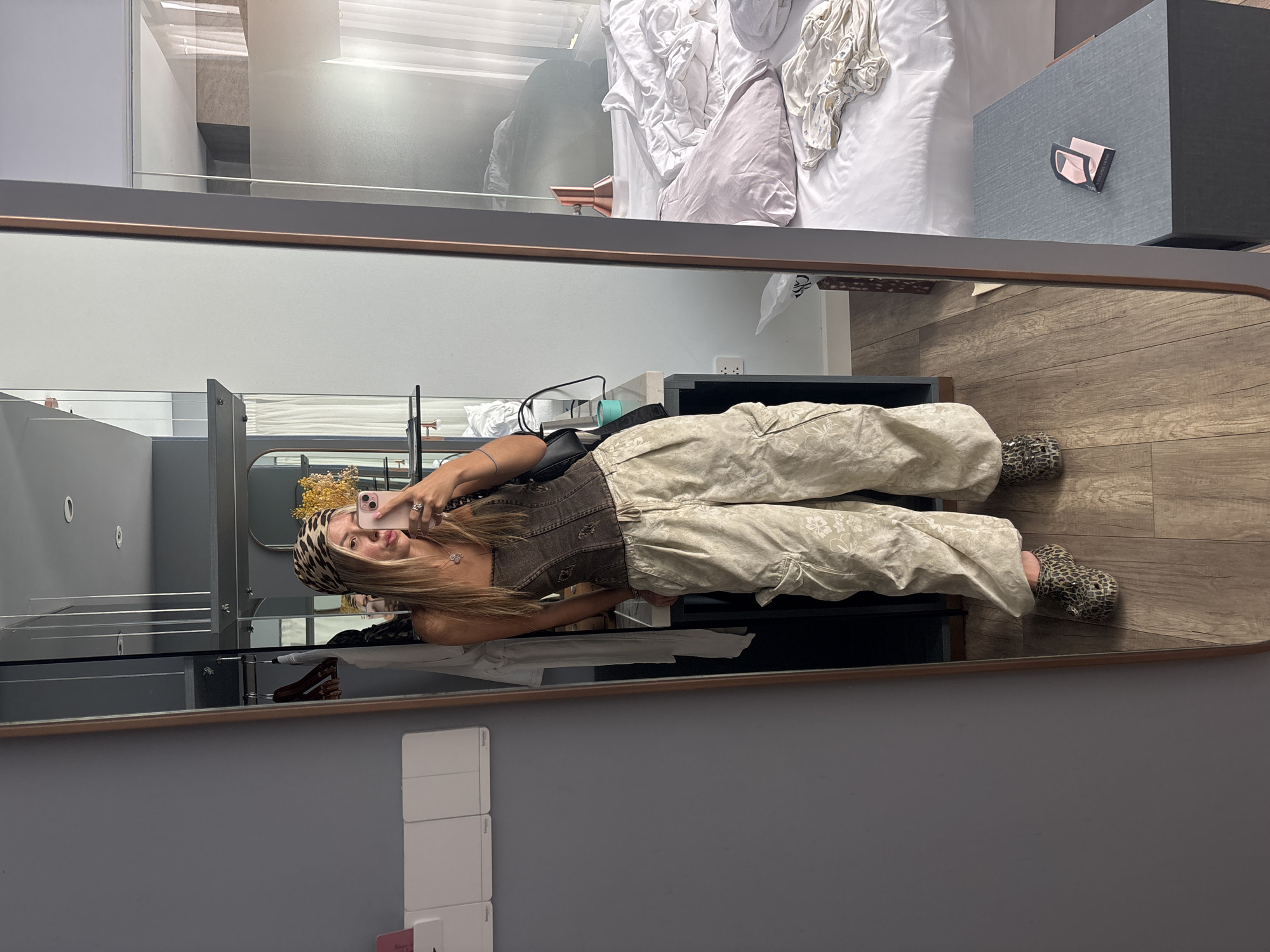 A young woman with long blonde hair and a leopard print headscarf takes a mirror selfie in a hotel room. She is wearing a brown corset top, baggy tan cargo pants with a floral print, and leopard print slip-on shoes. She holds her phone in her right hand. In the reflection, a messy bed with rumpled white sheets and a pink pillow is visible, along with a bedside table with a dark grey surface and a small item on top. The room has a modern aesthetic with grey walls and wooden flooring.