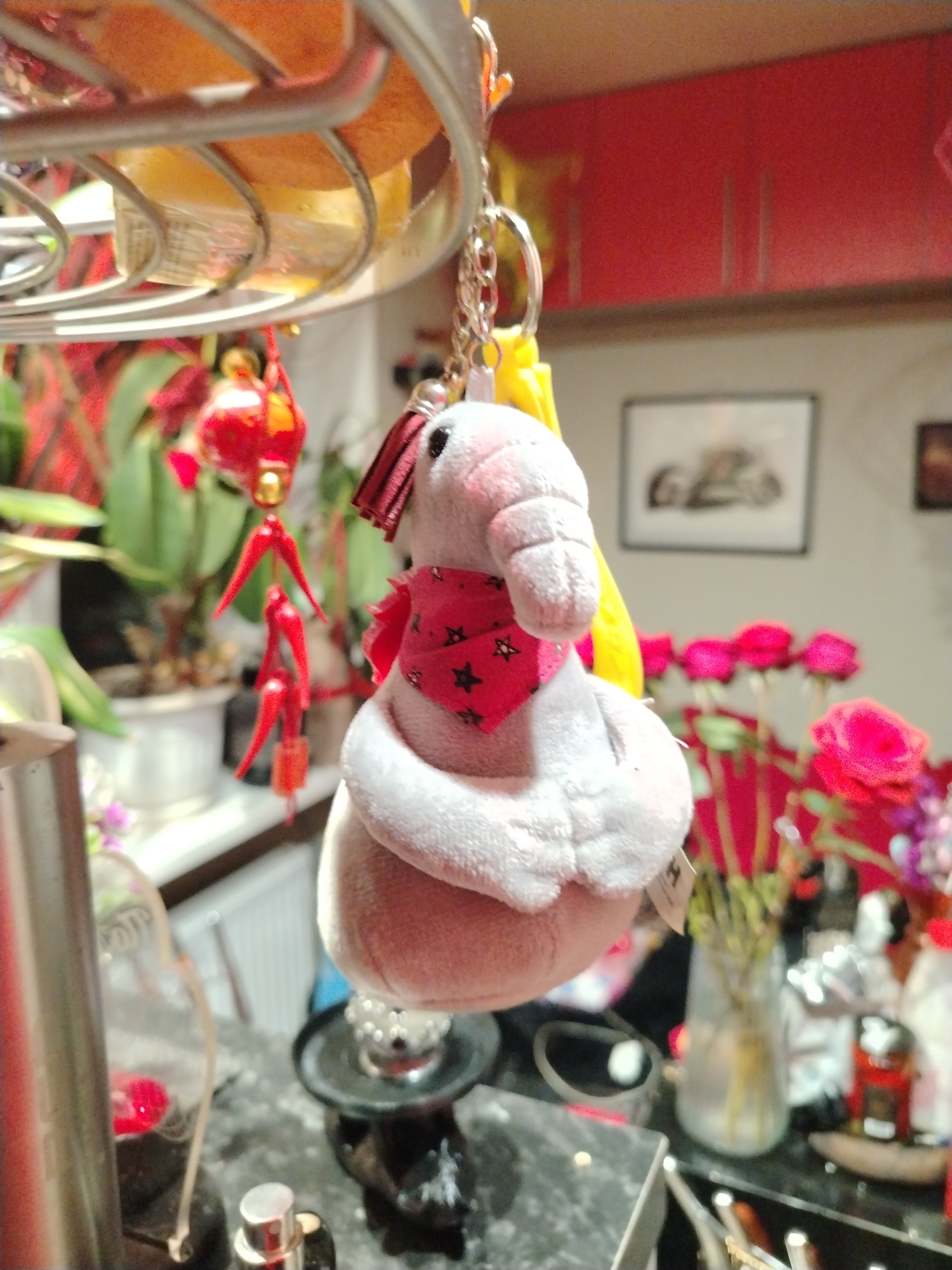 The image presents an indoor scene in what appears to be a domestic setting, possibly a kitchen or dining area. A plush, light gray toy animal with a red bandana is suspended from a silver-colored rack overhead. The background is filled with various items, including red cabinets, a framed picture on the wall, a vase containing red roses, and various decorative items. The foreground includes elements of a kitchen or dining setup, such as a black countertop and parts of what could be appliances or containers. The scene seems cluttered with personal items, suggesting a lived-in space. The location is given as Chaikovsky, Russia, but there is no visual evidence in the image to confirm this geographical location.