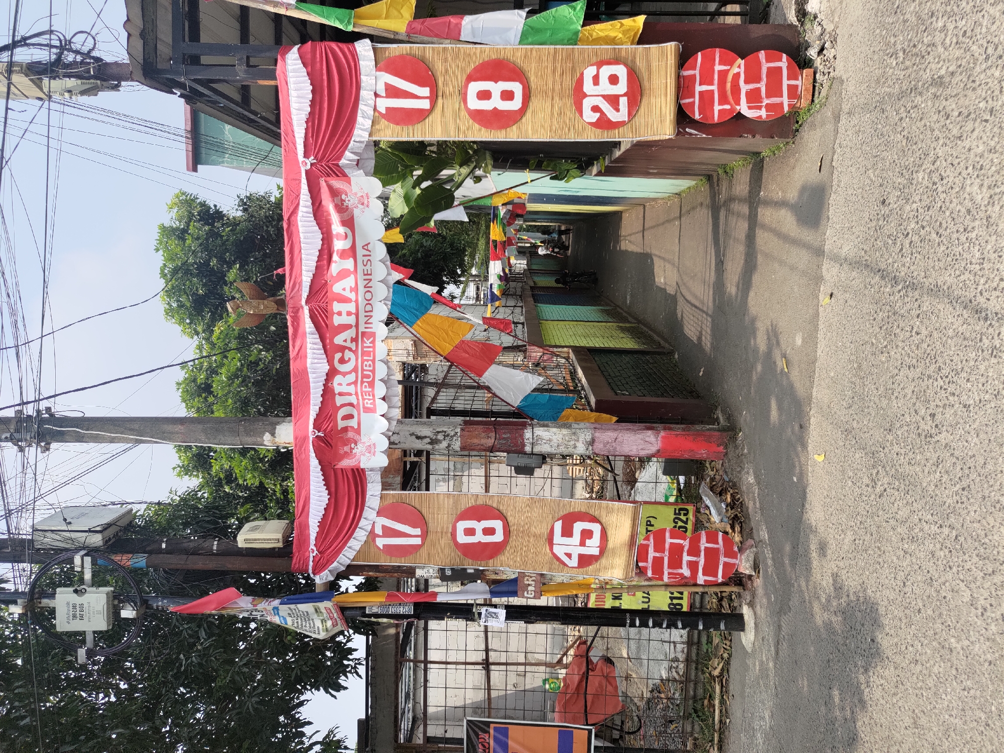 The image shows a street scene in Depok, Indonesia, with a decorated archway across a narrow alleyway. The archway is draped with red and white fabric, a national flag of Indonesia, and colorful pennant banners. A banner across the top of the arch reads "DIRGAHAYU REPUBLIK INDONESIA" (Long Live the Republic of Indonesia). To the left and right of the alley, vertical bamboo-like structures display large red circles with white numbers: "17", "8", and "45" on the left, and "17", "8", and "26" on the right. Below the numbers, there are circular decorations resembling red bricks with white lines. The alley itself is lined with colorful walls and leads to a dimly lit area where two people are visible, one on a motorbike. The street in front of the archway is paved and shows shadows from overhead wires and trees. Various electrical poles and wires are present in the background, along with some signage and a fence. The overall scene suggests a celebration of Indonesian Independence Day.