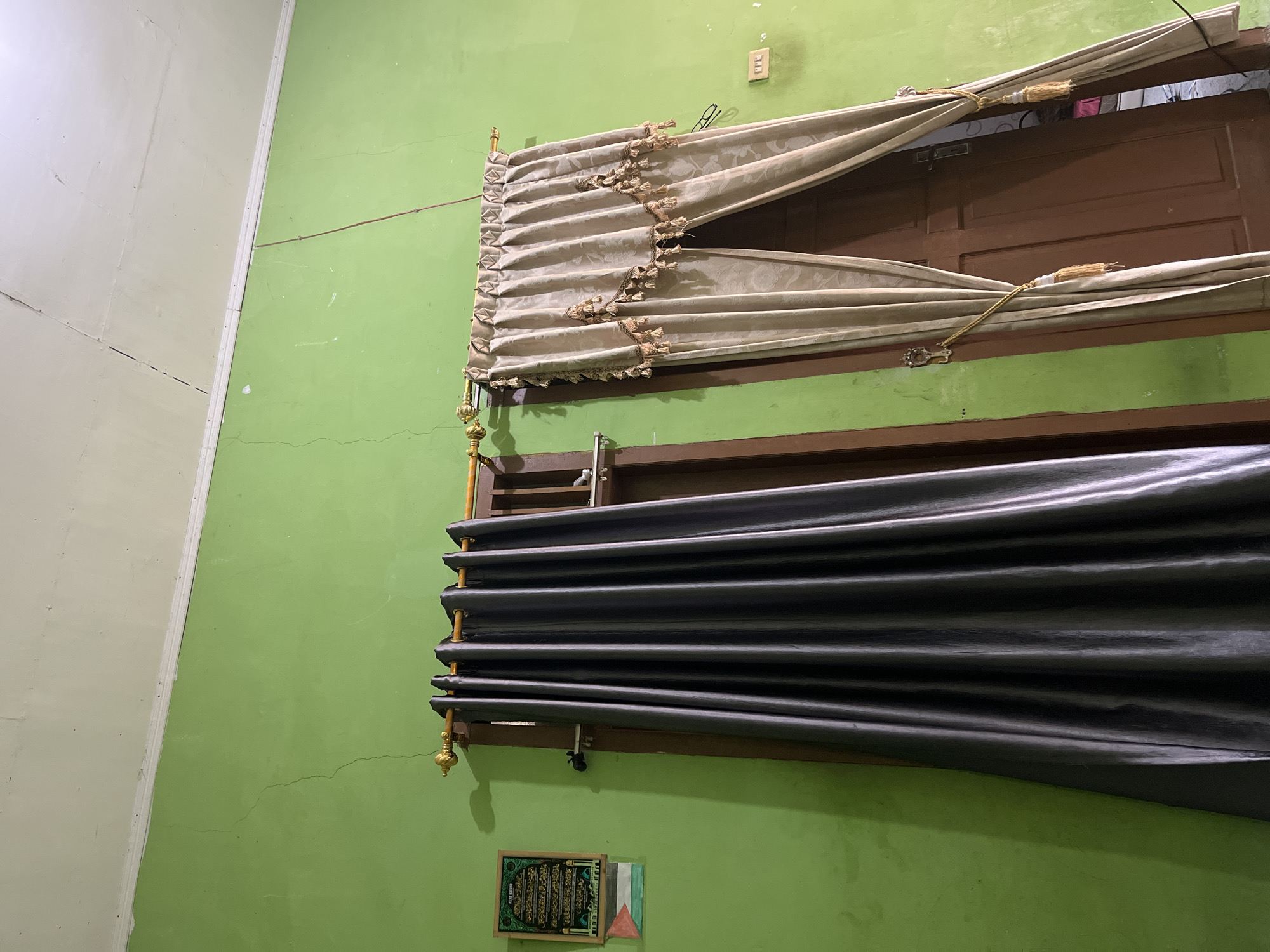The image depicts a room with bright green walls, likely a residential interior in Babahjurong, Indonesia. Two windows, draped with different curtains, are visible. The left window is covered by a dark gray, heavy curtain, while the right window is adorned with a lighter, beige curtain featuring decorative tassels. A golden curtain rod spans across both windows. A small framed picture, possibly religious text or art, hangs on the green wall to the left of the dark gray curtain, with a small flag beneath it. The ceiling appears to be white and shows signs of cracking. The floor is not visible. The scene is lit by artificial light, casting subtle shadows. The overall impression is of a simple, lived-in space.