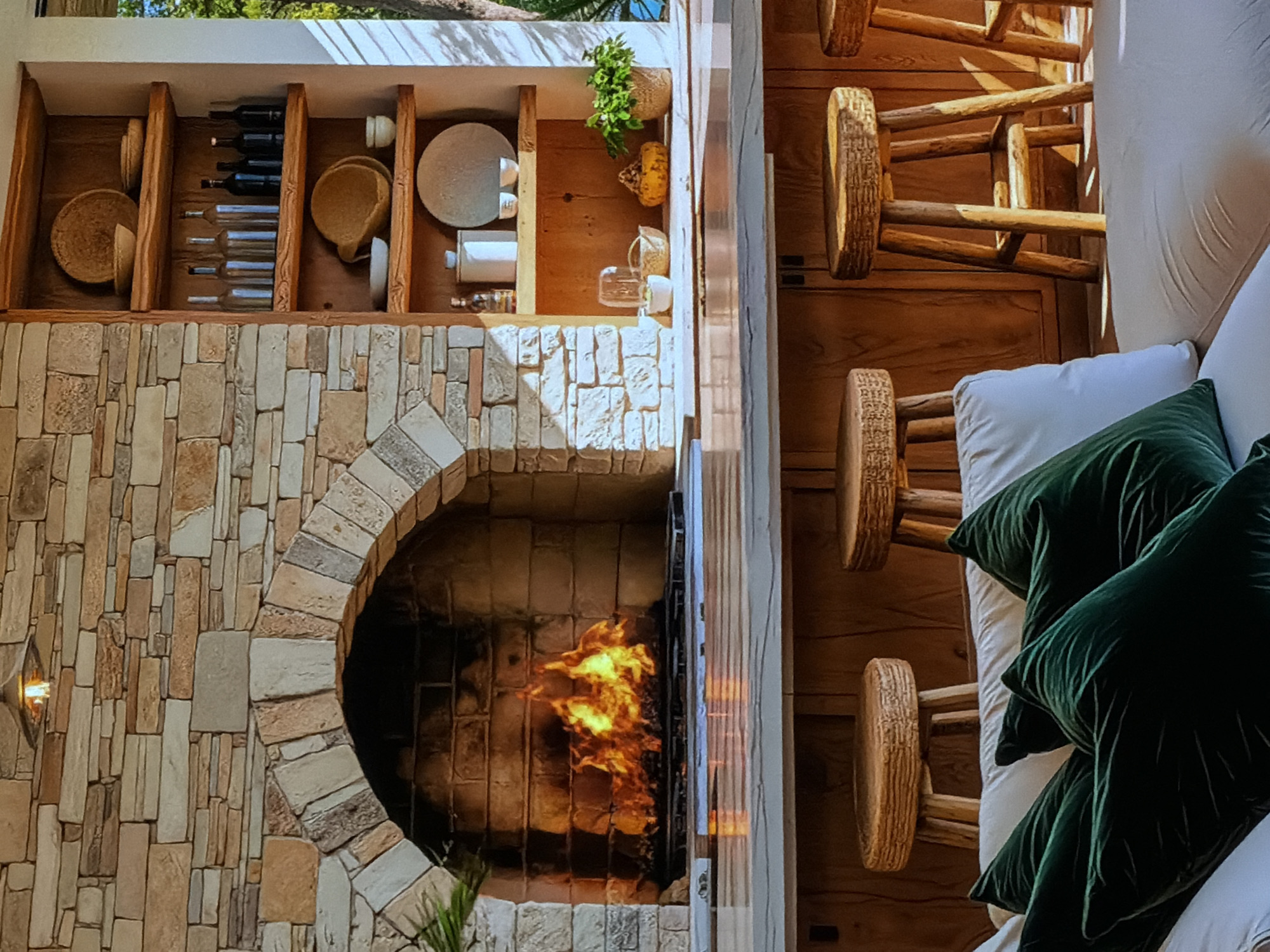 This media file captures a cozy and rustic indoor-outdoor living space, likely a lounge or bar area, set in Sofiivska Borshchahivka, Ukraine. No people are visible.
The focal point is a large, arched fireplace constructed from light-colored, rectangular stone blocks. A warm fire with bright yellow and orange flames burns inside, casting a glow. To the left of the fireplace, a small, warm light fixture is embedded in the stone wall.
Above the fireplace, a built-in wooden shelving unit serves as a display or bar area. It features vertical wooden dividers creating multiple compartments. The left section holds several woven baskets or bowls. The central section displays dark and clear bottles, possibly wine or spirits. The right section contains white ceramic dishes, a cylindrical container, and a small potted green plant. Further right on the shelf, more bottles, a glass, and a decorative gourd are visible.
To the right of the fireplace, a comfortable seating area is present, featuring a white sofa or bench adorned with large, plush dark green velvet throw pillows. In front of this seating, three rustic wooden stools with thick, round seats and sturdy, irregularly shaped legs suggest a bar or counter area, though the counter itself is not fully visible.
Natural light streams in from an opening at the top of the frame, illuminating the space and casting shadows. Green foliage, likely from trees, is visible through this opening, indicating an outdoor view or a semi-open structure. The overall atmosphere conveyed is one of warmth, comfort, and relaxation, with a blend of natural textures like stone, wood, and soft fabrics. No text is visible in the image.