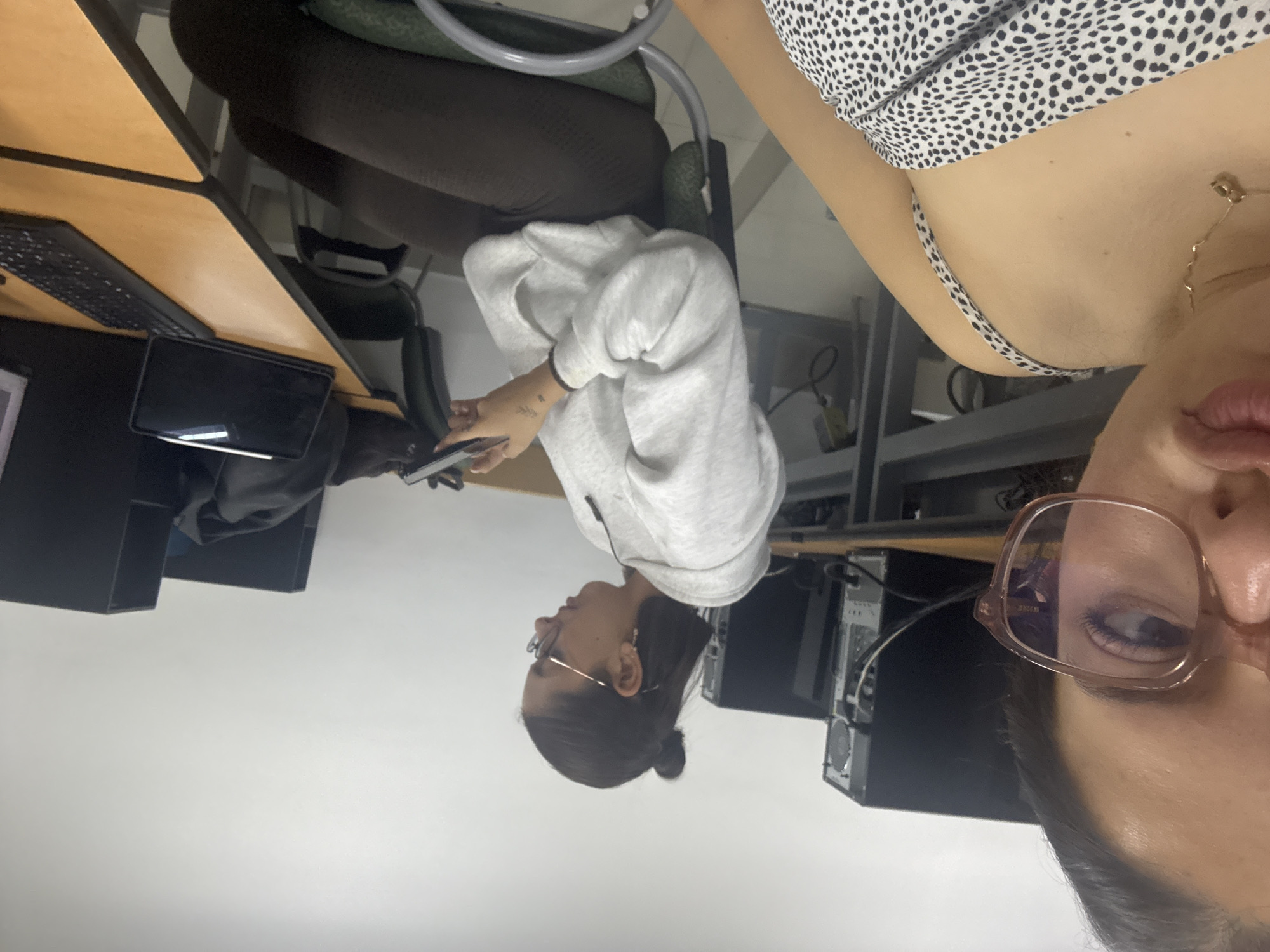The image, taken indoors, depicts two individuals in what appears to be a computer lab or office setting in San Andrés Cholula, Mexico. The perspective is from the immediate right of the frame, capturing a partial view of one person and a more complete view of another seated person.
In the foreground, on the right side, the face of a person is visible from the nose down, wearing light-framed glasses. Her lips are slightly parted, and she wears a gold chain necklace. Her top has a black and white spotted or animal print pattern. This individual appears to be holding the camera.
To the left, a second person is seated at a light-colored wooden desk with a dark trim. This individual wears a light grey hooded sweatshirt and dark pants or leggings. Her dark hair is pulled back, and she also wears glasses with a thin, possibly gold-toned, frame. She is looking to her left, seemingly engaged with a smartphone she holds in her left hand. A small, dark tattoo is visible on her right wrist.
The desk in front of the seated person holds a black keyboard and a dark tablet or another smartphone. In the background, behind the seated person, several dark computer towers are visible, connected by various cables, reinforcing the impression of a technical workspace. A dark office chair with a green cushion is positioned at the desk. The walls are light-colored, suggesting a well-lit indoor environment, likely daytime. No discernible text is visible in the frame.