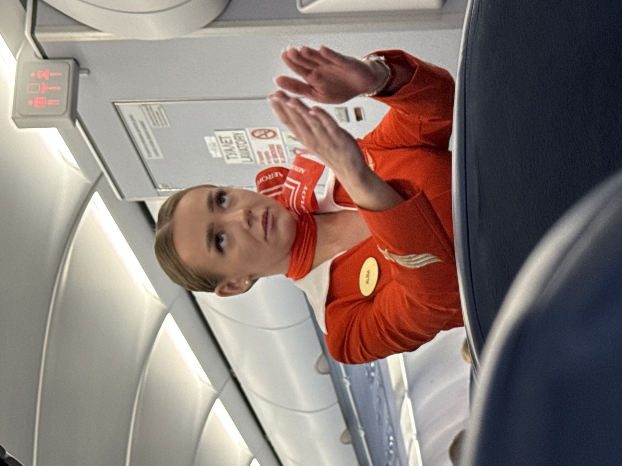 The image depicts an Aeroflot flight attendant wearing a red uniform with a white collar and red scarf. She has her hair pulled back and is looking upwards with her hands clasped together, as if demonstrating something. A name tag on her uniform reads "ALINA". In the background, there is a sign indicating "TOILET LAVATORY" with pictograms of a male and female restroom. The setting appears to be the interior of an airplane cabin, with curved white overhead panels and dark blue seats visible in the foreground. The presence of Aeroflot branding and the common interior of a passenger aircraft strongly suggest this photo was taken on an Aeroflot flight, likely at Khimki, Russia, which is home to Sheremetyevo International Airport, a major hub for Aeroflot.