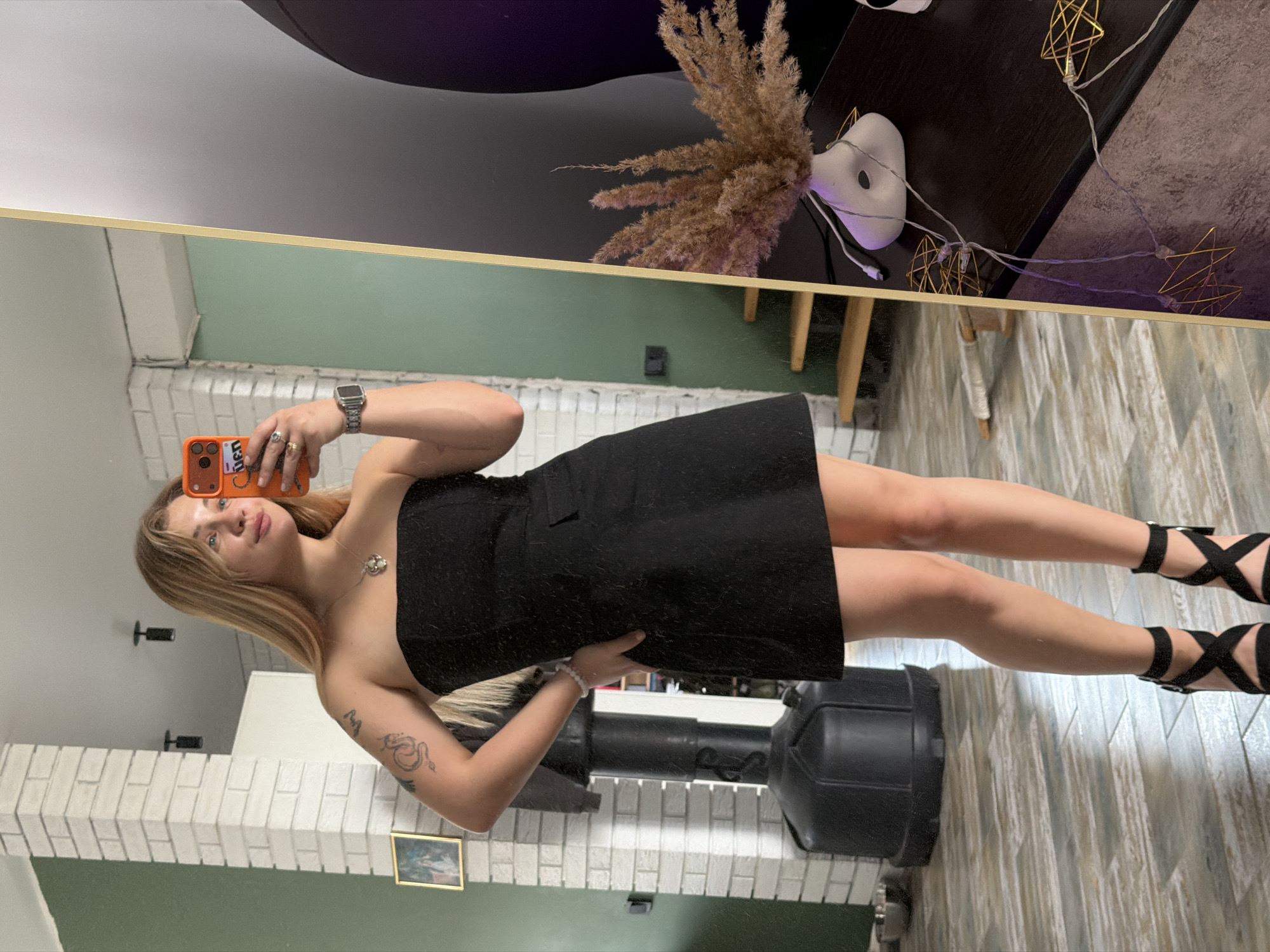 A woman stands in front of a mirror, holding a phone in her right hand and posing for a photo. She is wearing a black strapless dress and black high-heeled sandals with straps. Her hair is blonde. A watch is visible on her left wrist, and a tattoo is on her left arm. The background includes a white brick wall, green walls, and a framed picture. A black punching bag stands behind her. Reflected in the mirror is a dark table with decorations, including a vase of dried flowers and string lights. The location is likely indoors, in a room in Большая Шильна, Russia.