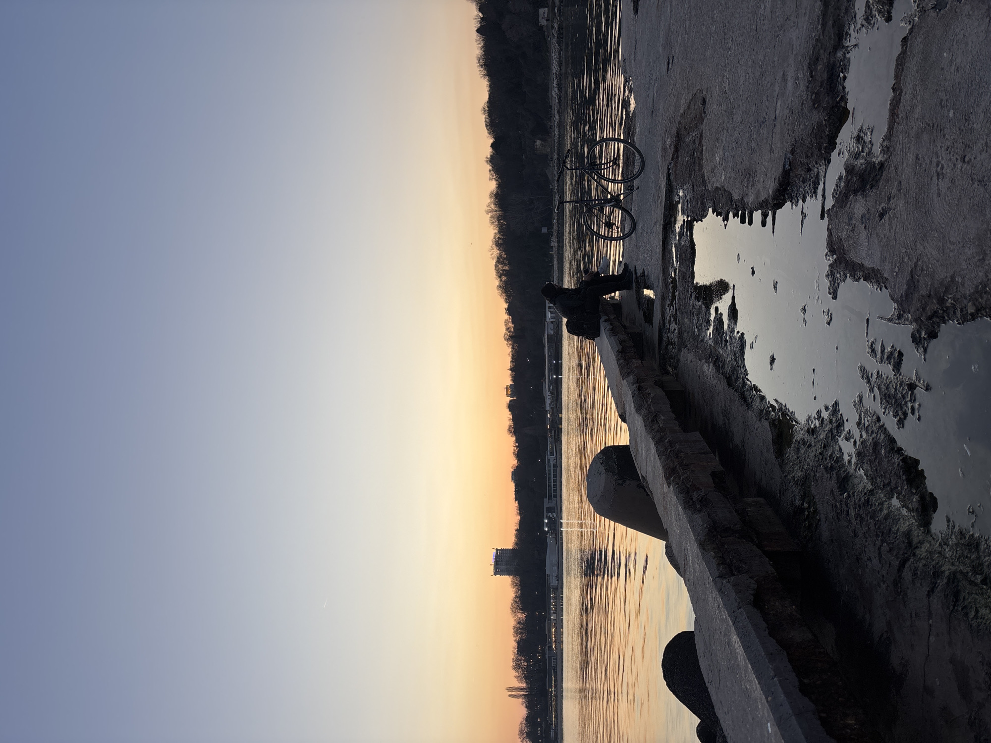 The image depicts a waterfront scene at sunset. A person is seated on a low concrete wall along the water's edge, with a bicycle parked nearby. The water reflects the orange and yellow hues of the setting sun. Beyond the water, there's a cityscape silhouette against the colorful sky. The foreground shows a concrete surface with puddles, which reflect the sky and surrounding elements. The location is unidentified.
