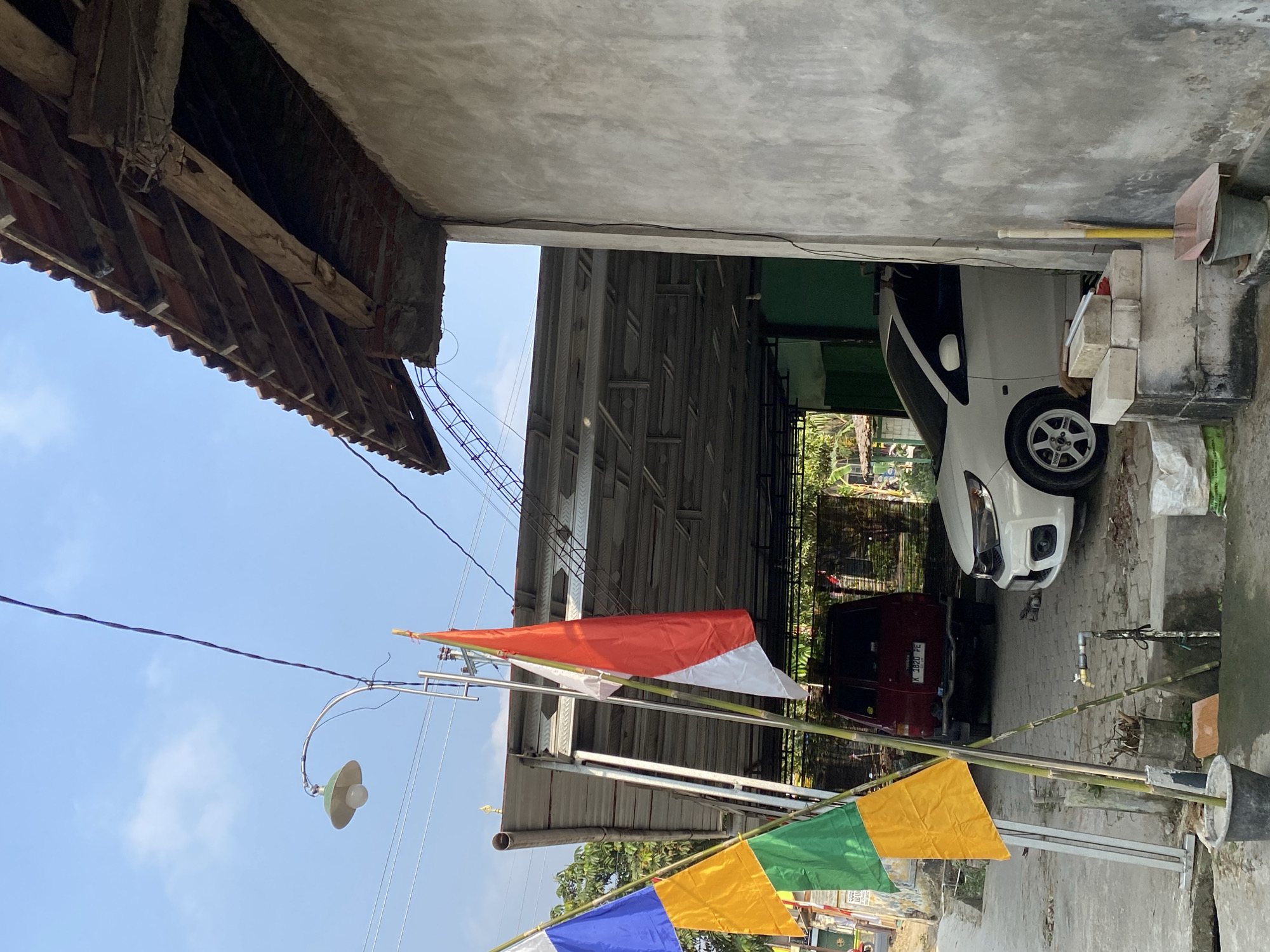 A medium shot captures an outdoor scene with a partially covered driveway or parking area. On the left, three flags are mounted on bamboo poles. The top flag is blue, the middle is yellow, and the bottom is green. Next to them, a tall pole supports the Indonesian flag, red on top and white on the bottom, and a white flag. A streetlamp with a green shade and a white bulb is visible in the upper left corner, attached to a metal pole. In the background, a red vehicle with license plate "K 1820 PE" is parked under a metal roof. To its right, a white car is parked under the same roof. The structure has a roof made of terracotta tiles and wooden beams. To the right of the scene, a rough concrete wall of a building occupies a significant portion of the frame, with electrical wires running along it. A simple faucet with a hose is mounted on a pipe near the ground in front of the white car. Bricks and bags of material are stacked near the building's wall. The sky above is clear blue with a few wispy clouds. The ground appears to be paved with interlocking concrete blocks.