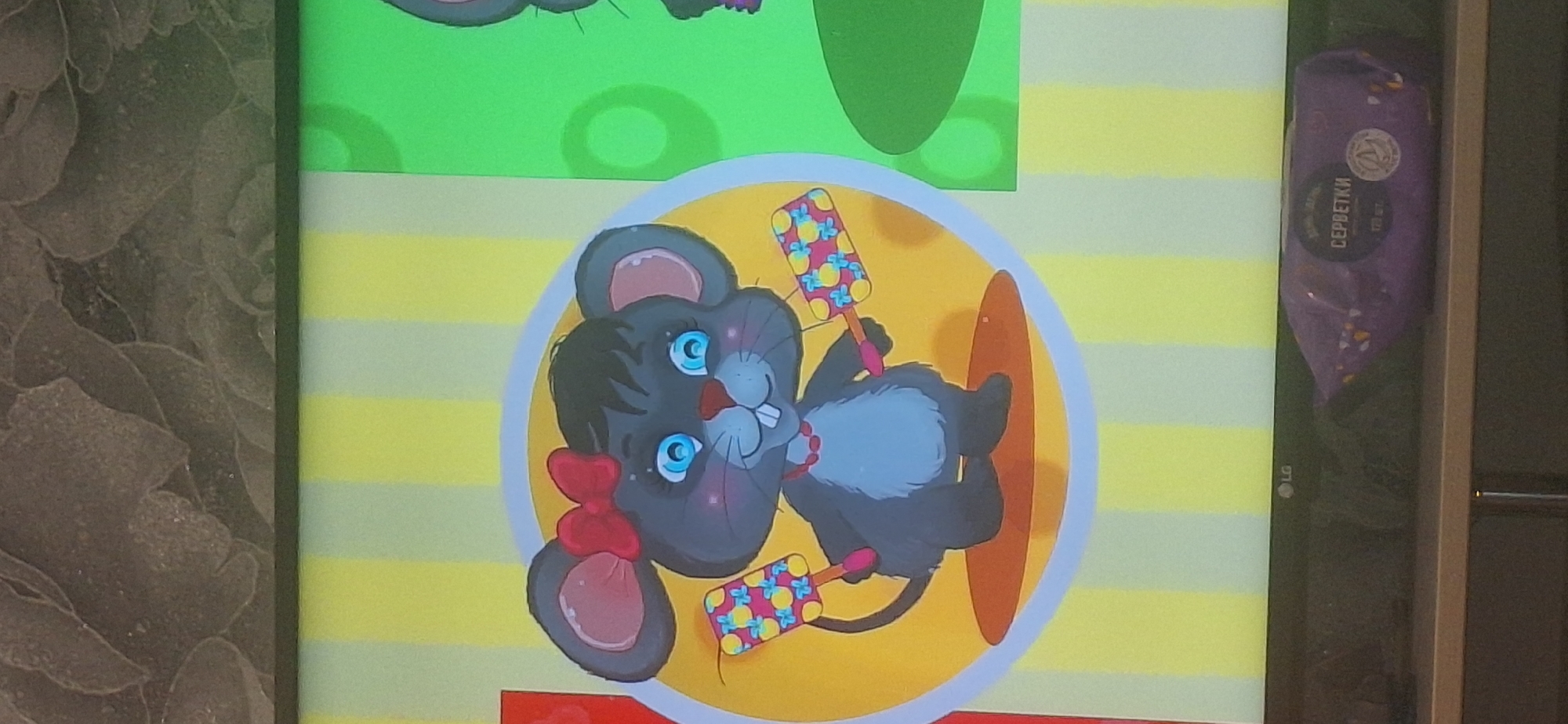 The image shows a cartoon mouse with a red bow, holding two colorful toys. The mouse is gray with pink ears, blue eyes, and a red necklace. It has two front teeth and is smiling. The background consists of vertical stripes of yellow and light green. To the right of the mouse, another cartoon character, partially visible, appears to be a mouse with pink shoes. Below the cartoon, a portion of a television screen with the LG logo is visible, and on the shelf beneath the television, a purple package of wet wipes is present. The image does not contain any information that would indicate its location in Bila Tserkva, Ukraine.