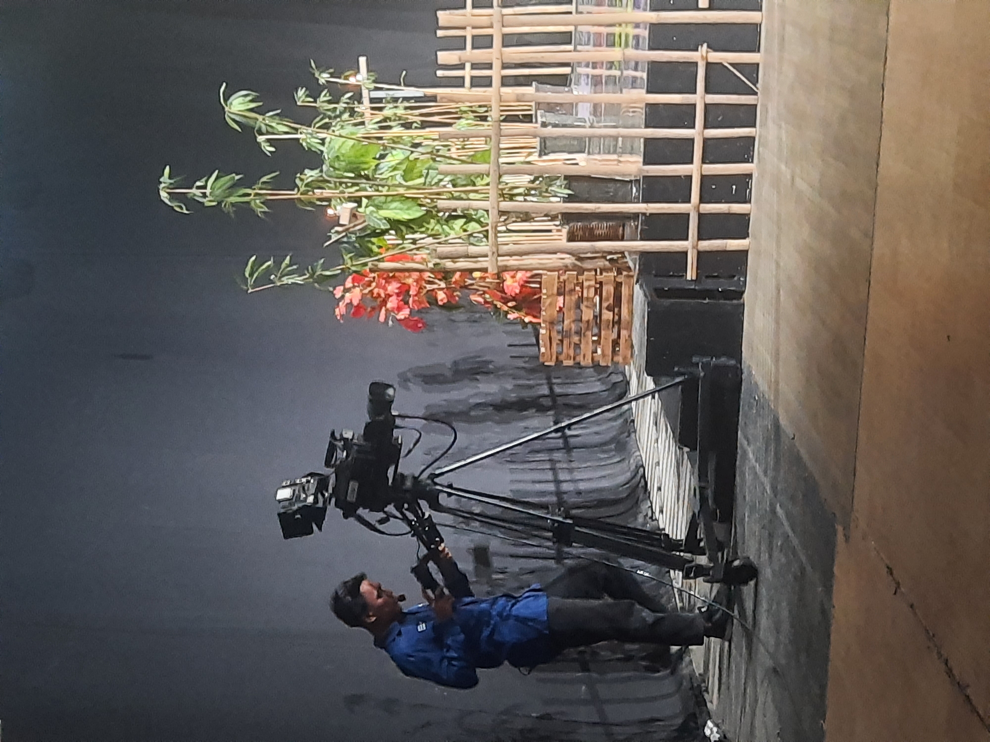 A man in a blue shirt and black pants is operating a professional video camera on a tripod. He is looking down at a device he is holding in his hands, possibly a remote or monitor. The camera is positioned on a stage with a dark backdrop. To the right of the camera, there are several potted plants, some with red foliage and others with green leaves and thin stalks, arranged behind a bamboo-like fence structure. There are also wooden crates and black boxes on the stage. The overall lighting suggests an indoor setting, possibly a studio or performance space. The scene appears to be a setup for recording or filming an event. The image does not contain enough information to determine the specific location in Bandung City, Indonesia.
