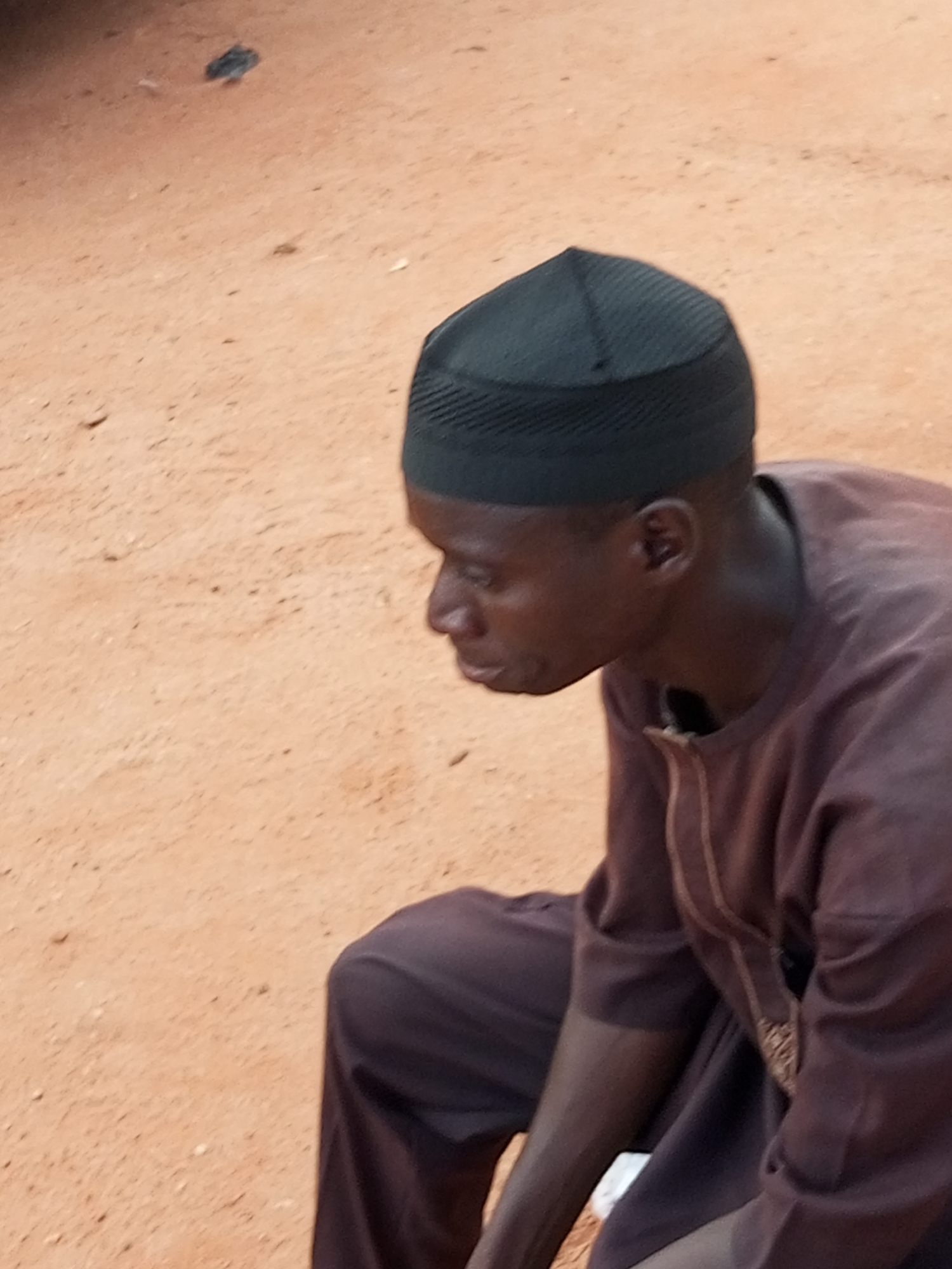 A man wearing a dark brown traditional outfit and a black kufi cap is sitting on the ground. He is looking down and to his left. The ground is sandy and appears to be an outdoor area. The image is taken from a medium distance, focusing on the man's upper body and head. The lighting suggests it is daytime, with soft shadows. The overall scene is simple and still, with no other individuals or prominent objects visible. The environment appears to be unpaved, possibly a street or an open area in Gombe, Nigeria.