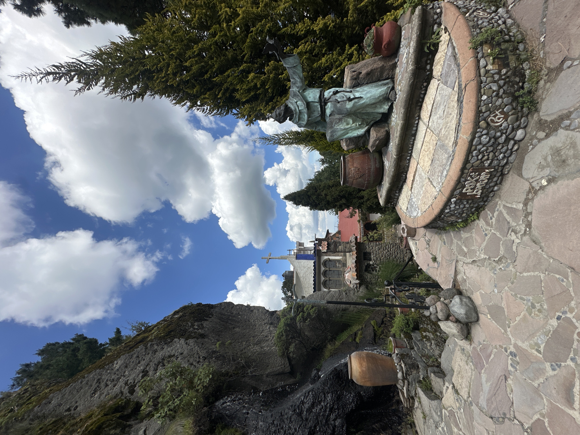 The image depicts a scene at a spiritual site in San Mateo Ozolco, Mexico. In the foreground, a pathway paved with irregular stones and pebbles leads towards a semicircular stone structure. Seated on this structure is a bronze statue of a robed figure, possibly a saint or monk, holding two birds in its outstretched hand. The statue is weathered and greenish, suggesting age and exposure to the elements. To the left of the pathway, a large, rustic clay pot rests near a rock face with a small waterfall cascading down it. Behind the pathway and the statue, buildings with red tiled roofs and stone construction are visible, one of which has a cross atop its roof. Lush green trees, including a tall conifer on the right, frame the scene, and a dramatic sky with white cumulus clouds adds to the atmosphere. The overall impression is one of tranquility and spiritual contemplation.