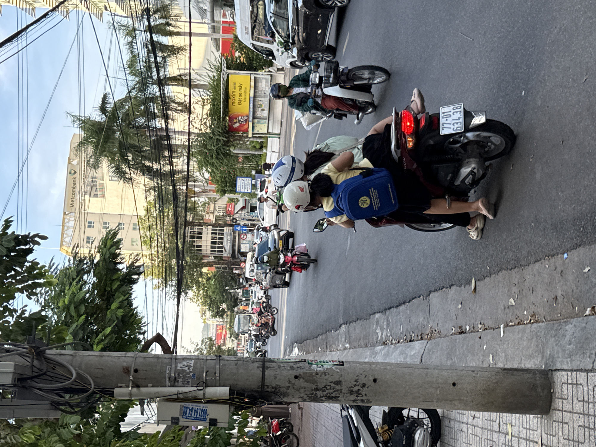 In Nha Trang, Vietnam, a city street is depicted with a moderate amount of traffic. A concrete utility pole stands prominently in the foreground on the left, with numerous electrical wires crisscrossing overhead. Beyond the pole, a sidewalk with a tiled pattern is visible, bordered by a concrete curb. The street itself is paved with asphalt and has several vehicles, primarily motorcycles, in motion. On the right side of the street, there are buildings with signs, including one for "Vietcombank" and another yellow advertisement for "maxim." In the center of the image, two individuals are riding a red scooter, with the person in the back wearing a blue backpack. Another scooter with a person wearing a green jacket and a mask is to the right, carrying a large white bag. A dark-colored car is also present in the traffic. The sky is partly cloudy, and palm trees are visible among the buildings, indicating a tropical or subtropical urban environment. The scene captures a typical street view in a bustling Vietnamese city, characterized by its active street life and prevalent use of motorcycles.