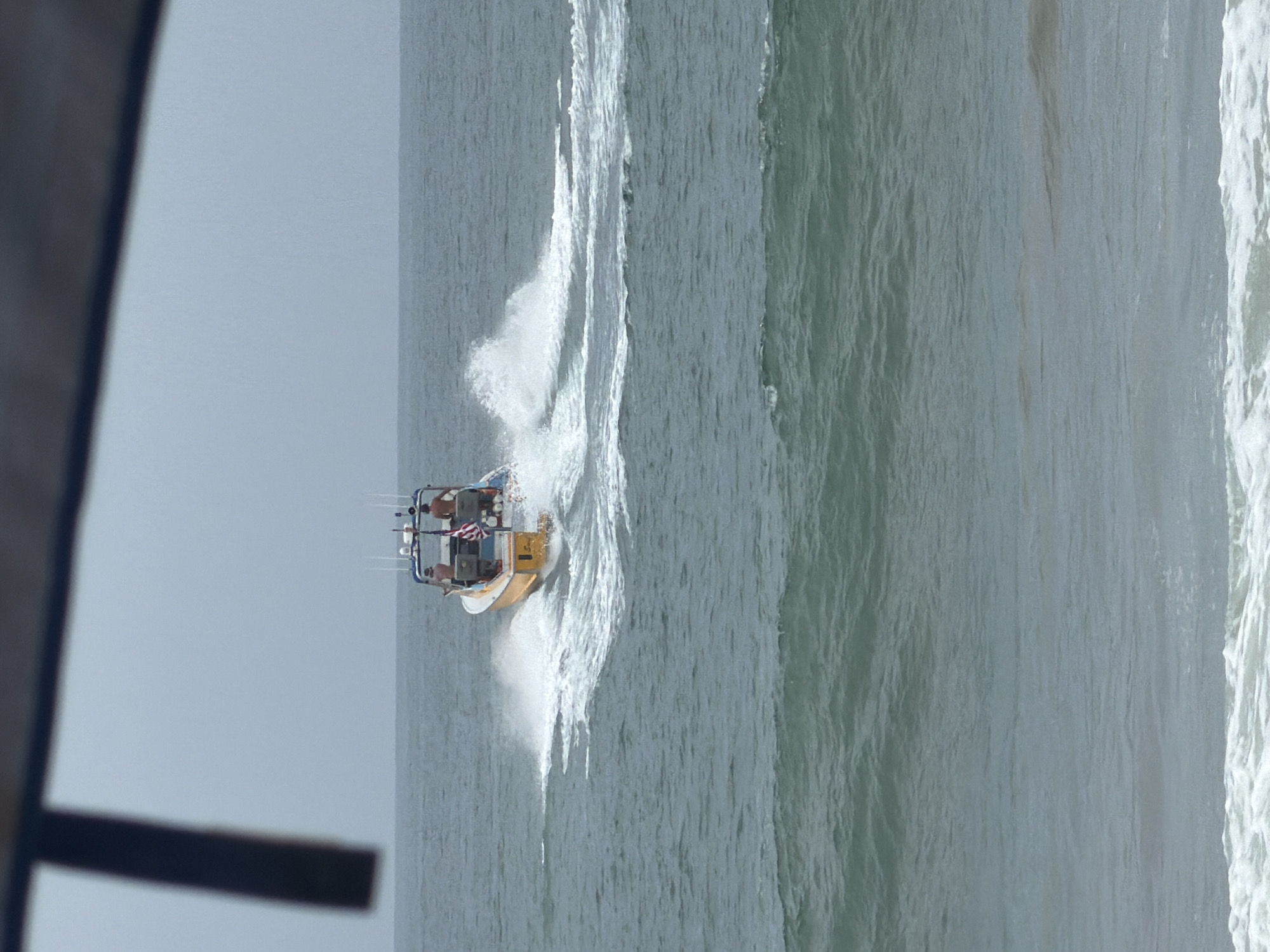 An offshore shot captures a yellow and white boat with two individuals on board, heading away from the viewer. The boat is leaving a white spray wake behind it, indicating movement through the water. The ocean appears choppy with moderate waves, and the sky is overcast and hazy, suggesting cloudy weather. The foreground of the image is dominated by the ocean, with the horizon line barely visible in the distance, separating the sea from the pale sky. The presence of a dark, angled object in the upper left corner of the frame suggests the photo was taken from an elevated vantage point, possibly from a structure or another vessel. The overall scene depicts a marine environment with a boat actively navigating the sea.