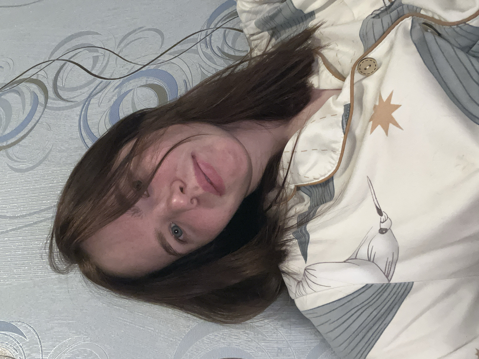 A young individual, likely female, with long brown hair and blue-grey eyes, is depicted lying down and gazing directly at the camera. Their head is positioned towards the upper-left of the frame (assuming standard orientation), with their body extending diagonally. A subtle, soft smile plays on their lips, suggesting a relaxed and slightly playful or curious expression. Their hair is casually arranged, framing their face.
The person is dressed in light-colored sleepwear, possibly pajamas, which features brown piping along the edges and a round, light brown button near the neckline. The fabric of the sleepwear is patterned with stylized motifs, including abstract blue-grey shapes, a distinct light brown star, and an illustration that appears to be a graceful, stylized bird or crane.
The background consists of a surface, likely a wall or bed, covered with light-toned material, possibly wallpaper or bedding. This material displays an intricate pattern of swirling lines and organic shapes in soft shades of light blue, grey, and brown. The setting suggests an indoor environment, consistent with a private residence in Chaikovsky, Russia. The lighting is soft and even, indicating either daytime indoors or a well-lit interior. There is no visible text present in the image.
