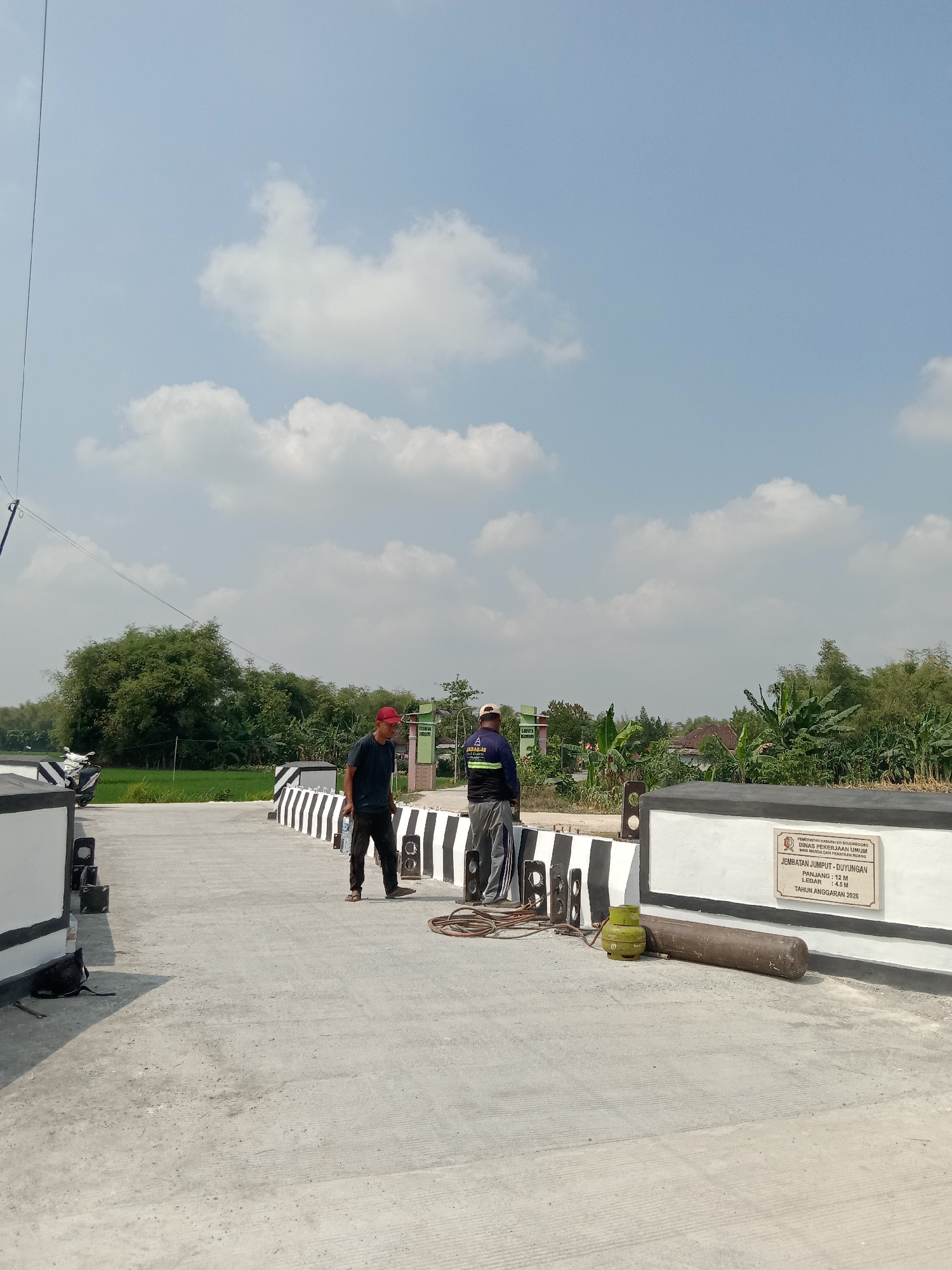 Two men stand on a concrete bridge under a partly cloudy sky in Semen Kidul, Indonesia. The man on the left wears a dark blue t-shirt, dark pants, and a red baseball cap. He holds a bottle of water and looks toward the right. The man on the right wears a dark blue vest over a long-sleeved shirt, grey pants, and a beige baseball cap. He faces away from the camera, looking toward a sign mounted on the bridge railing. The bridge has white and black striped barriers. In the foreground, a green gas cylinder and a brown, cylindrical object lie on the ground next to coiled rope. A motorcycle is parked on the left side of the bridge, and lush green vegetation, including trees and banana plants, surrounds the bridge and fields. A sign on the bridge railing reads, "PEMERINTAH KABUPATEN BOGOR DINAS PEKERJAAN UMUM BINA MARGA DAN PERAIRAN RUANG JEMBATAN JUMPUT - DUYUNGAN PANJANG : 12 M LEBAR : 4,5 M TAHUN ANGGARAN 2026."