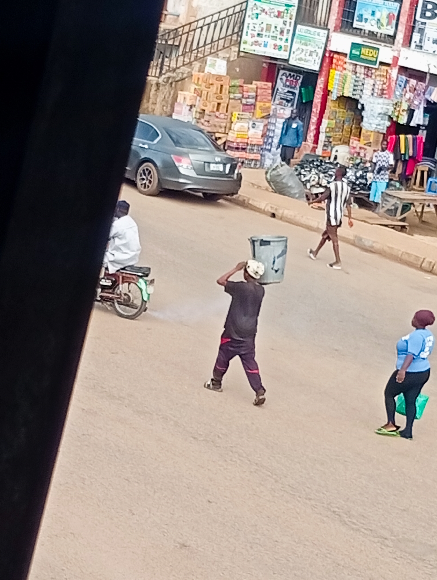 The image depicts a street scene in Bukuru, Nigeria, likely during daytime. In the foreground, a young man wearing a dark t-shirt and purple trousers carries a large, grey cylindrical bin on his shoulder, facing away from the viewer. To his right, a woman in a light blue t-shirt and black leggings stands facing left, holding a green plastic bag. Further down the street, another man in a black and white striped shirt and light blue shorts walks away from the viewer. A motorcycle with a rider dressed in a white garment is visible on the left side of the image, producing a trail of smoke. In the background, a dark-colored sedan is parked on the right side of the road. The street is lined with shops and market stalls displaying various goods, with stacked boxes and hanging items visible. The ground appears to be a sandy or dirt road.