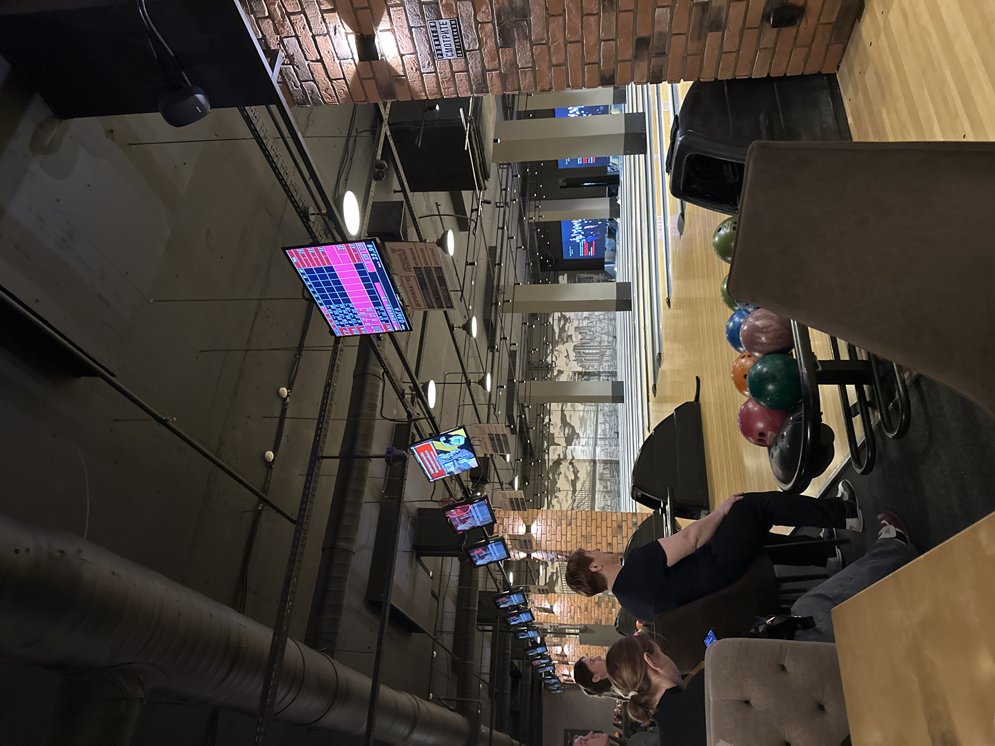 A bowling alley in Izhevsk, Russia is shown from a low-angle, indoor perspective. The venue features multiple lanes with overhead scoring screens. On the right, a group of bowling balls is visible along with seated individuals, one of whom is poised to bowl. Overhead, the venue has a network of pipes, lighting fixtures, and numerous monitors. Exposed brickwork accents the upper walls. The scene is illuminated by ambient lighting.