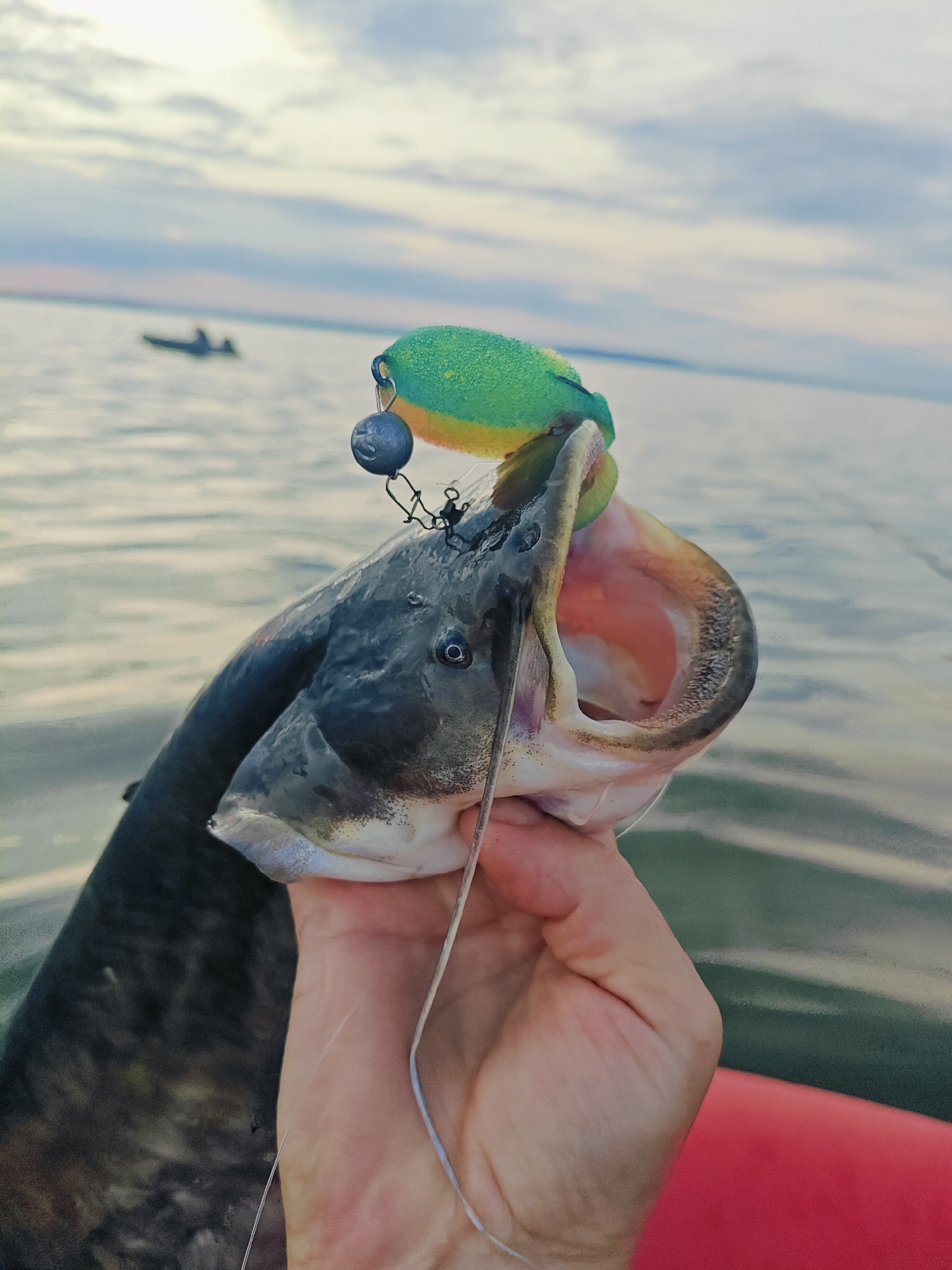 A person's hand holds a large catfish, its mouth agape, revealing pink flesh and sharp teeth. A green and yellow fishing lure with a small, dark gray weight is lodged in the fish's mouth. The fish is dark gray and appears to be recently caught, with a fishing line visible. The background shows a calm body of water, likely a reservoir or large river, with a cloudy sky overhead. In the distance, a small boat with at least one person can be seen. The foreground is occupied by a portion of what appears to be a boat or raft in a red color. The image was taken on the Kama River in Chaykovsky, Russia.
