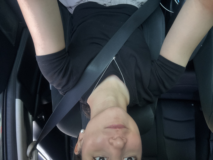 The image depicts an individual, presumed female, seated in the driver's position inside a vehicle. The upper torso, arms, and lower half of the face are visible. The individual has a light to medium skin tone, dark eyes, and a slight smile. A white pearl earring is visible on the right ear. The individual is wearing a black short-sleeved t-shirt, a thin silver chain necklace with a small pendant, and a light-colored lower garment, partially visible below the waist. A dark-colored seatbelt is correctly fastened across the individual's chest and lap. The vehicle's interior features dark upholstery, consistent with car seats and interior trim. The back of a rear passenger seat is visible behind the individual. Lighting conditions suggest daytime. While the prompt specifies the location as Santa Catarina, Mexico, the visual information within the image does not provide any discernible cues to confirm this geographical detail or a specific location within the city.
